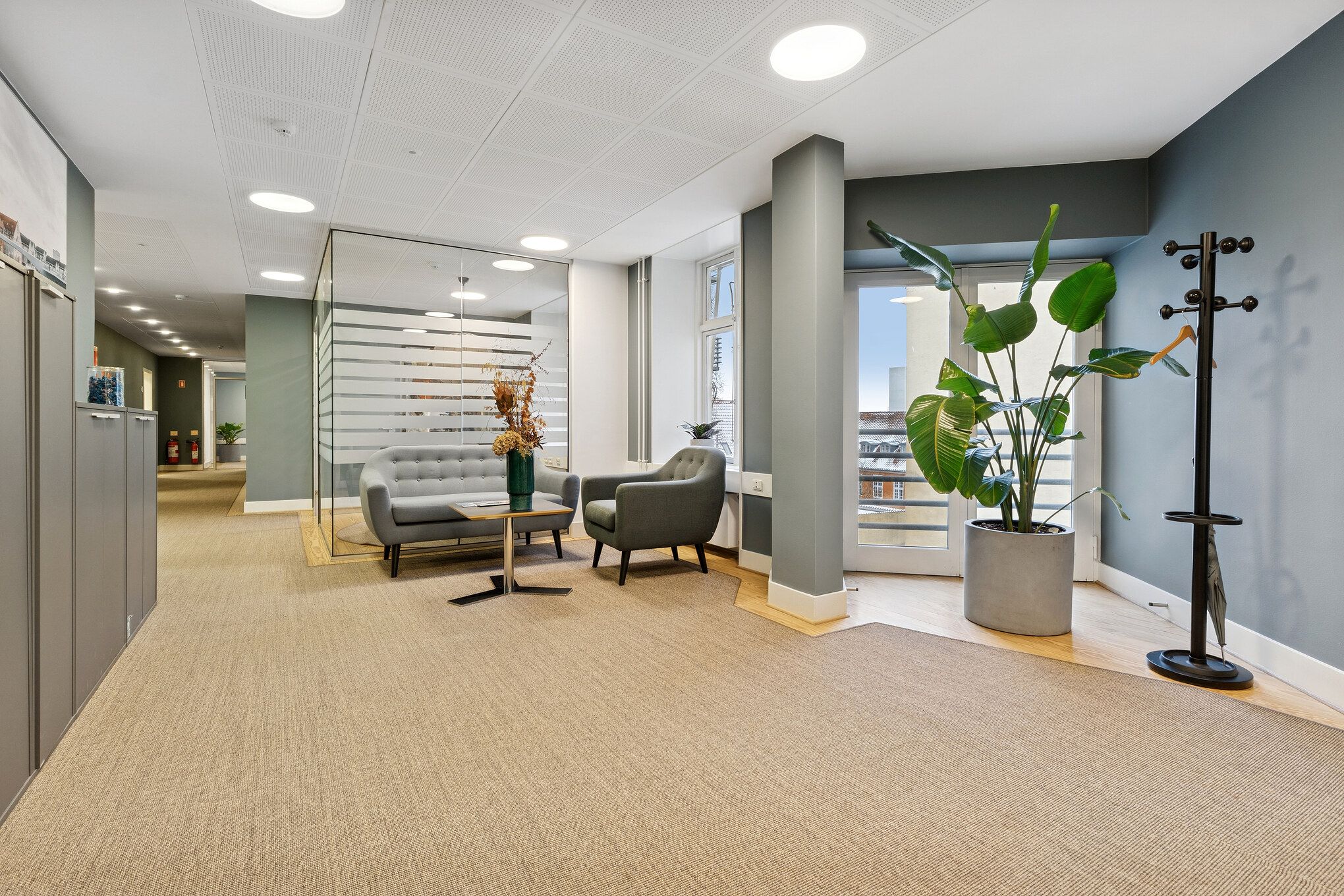 Kontorområde med sofaer og bord, indretning med plante og hængeklæder, moderne loftslampe og møbler, trægulv og lyse ruder