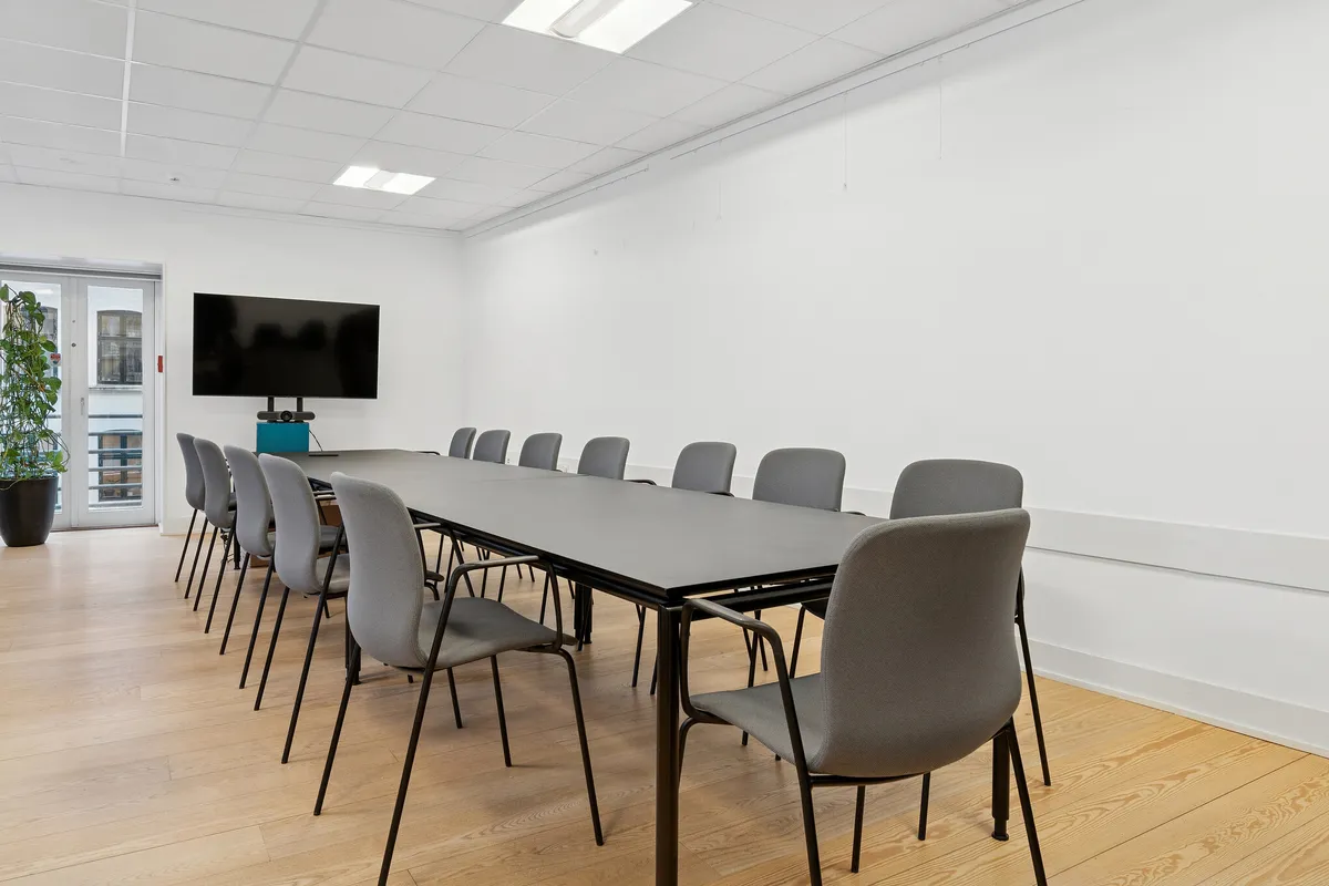 Kontor med lyse møbler, lysegule vægge, trægulv, tv, gruppe mødebord og stole samt plante i potte ved indgangen