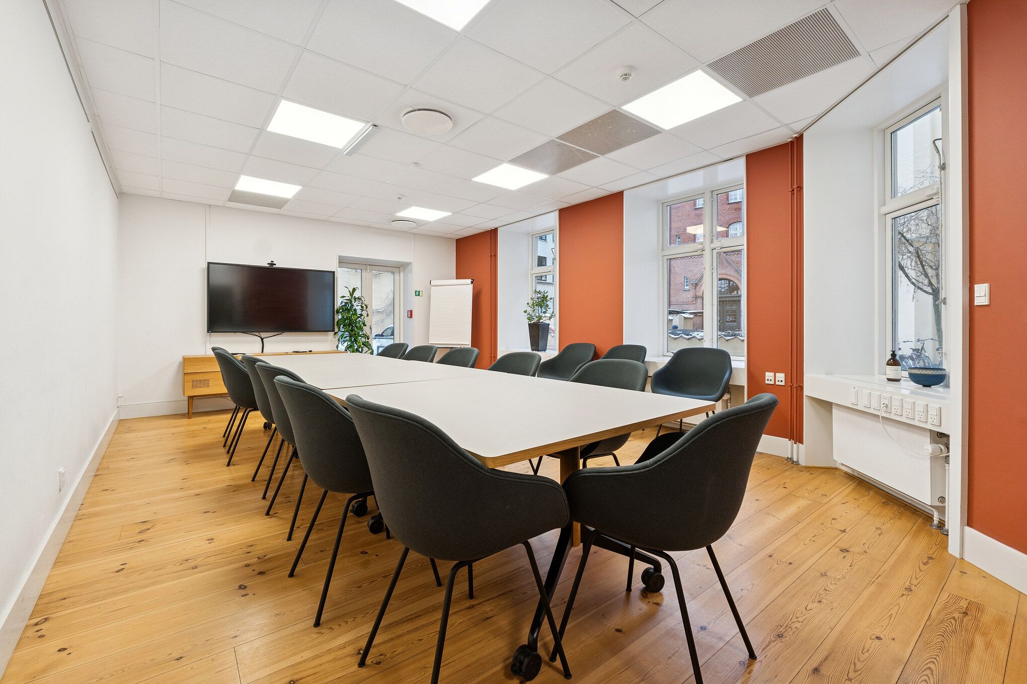 Kontorrum med højt til loftet, lyse loftslampe, møbler, plante, whiteboard, TV, udendørsudsigt og trægulv i et velfungerende flerbrugerhus i Københavns historiske kvarter.