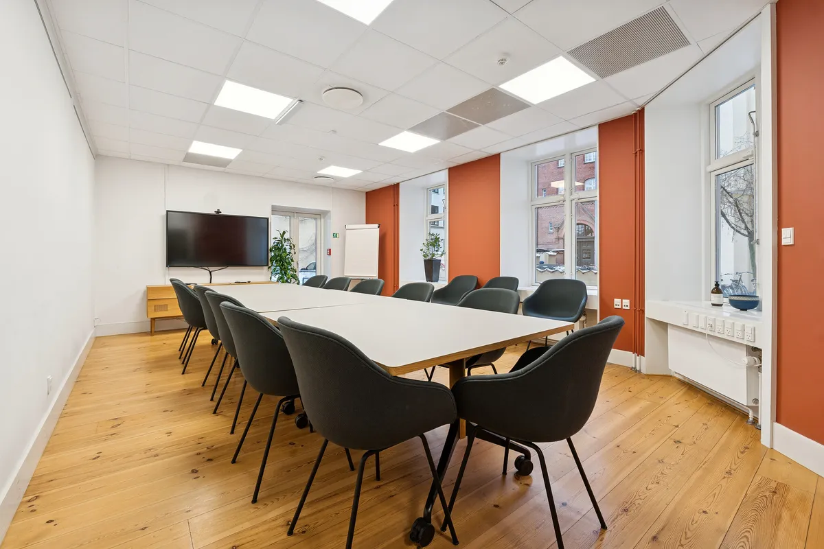 Kontorrum med højt til loftet, lyse loftslampe, møbler, plante, whiteboard, TV, udendørsudsigt og trægulv i et velfungerende flerbrugerhus i Københavns historiske kvarter.
