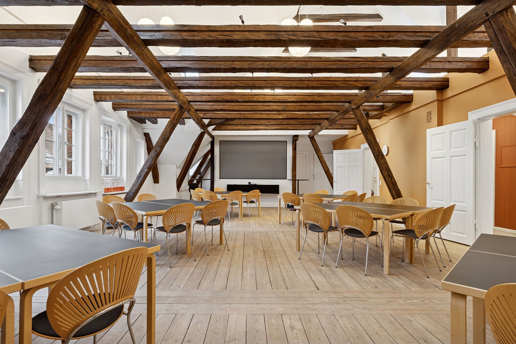 Kontorrum med møbler og træloft i et historisk kvarter, moderne møbler og tekstiler, fire store vinduer og en stor skærm, åbne døre og lys pære på loftet