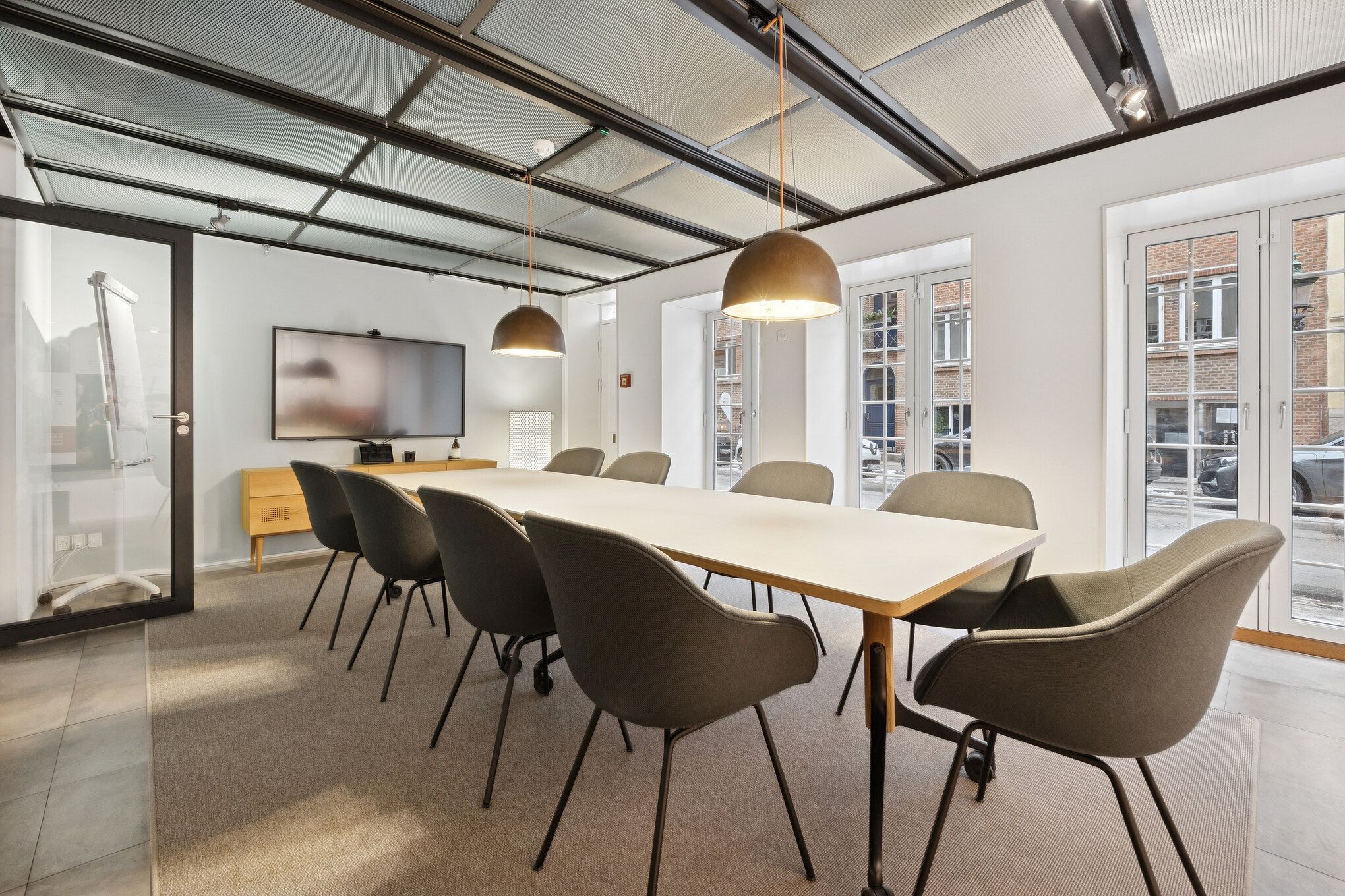 Kontorrum med moderne møbler, hvidt loft, træbord, moderne stolpelysekroner og tv-skærm samt glasdøre og vinduer i baggrunden
