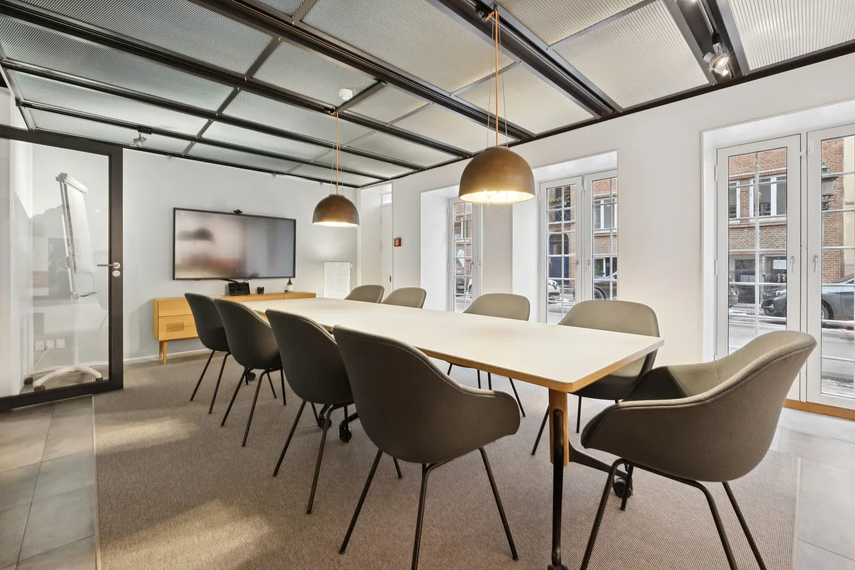 Kontorrum med moderne møbler, hvidt loft, træbord, moderne stolpelysekroner og tv-skærm samt glasdøre og vinduer i baggrunden