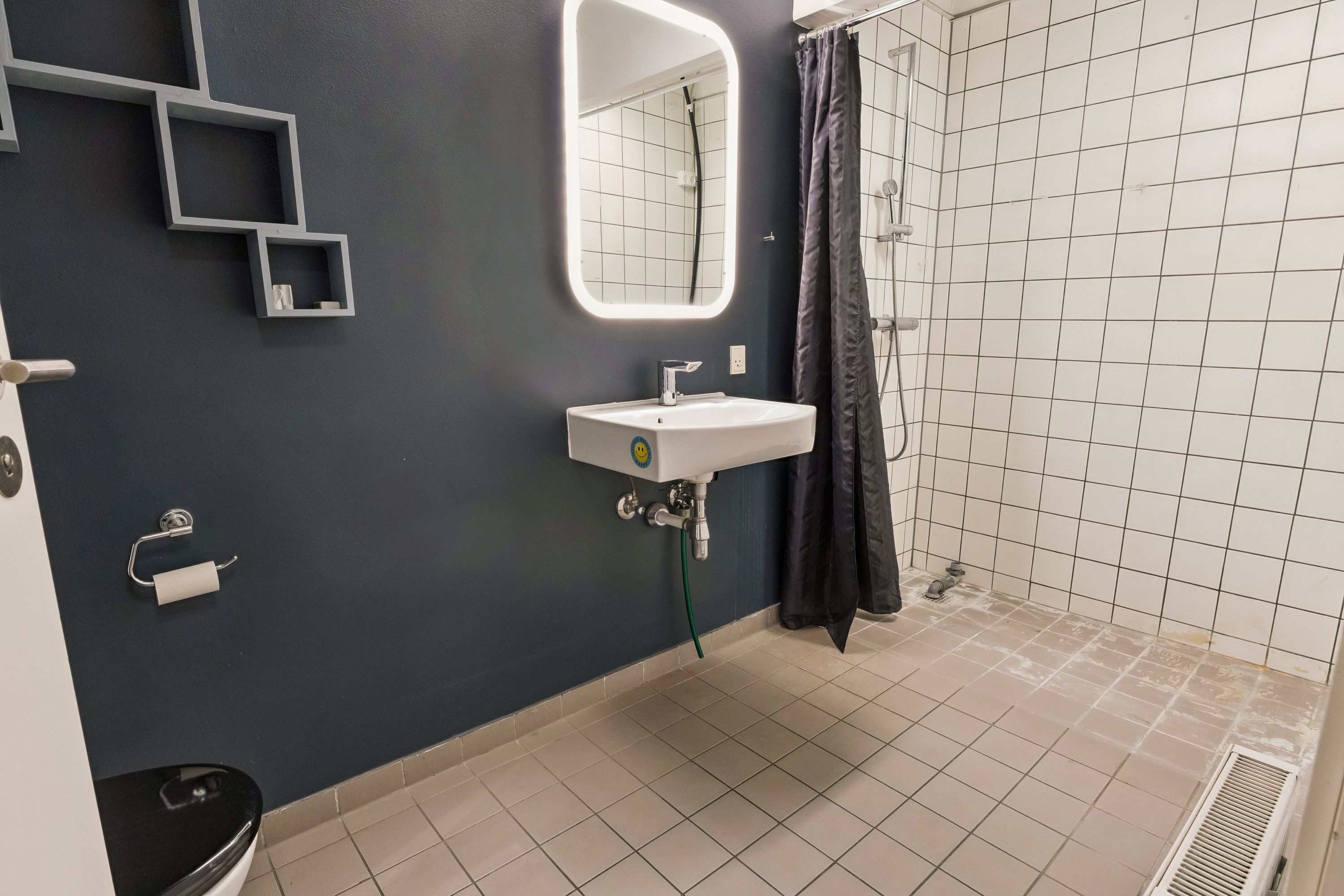 Et lille badeværelse med hvid flise og sort toilet på gulvet, lysegrå væg med et højt spejl og tre rummelige hylder, lysegrå sæbeholder og hvidt toiletpapir