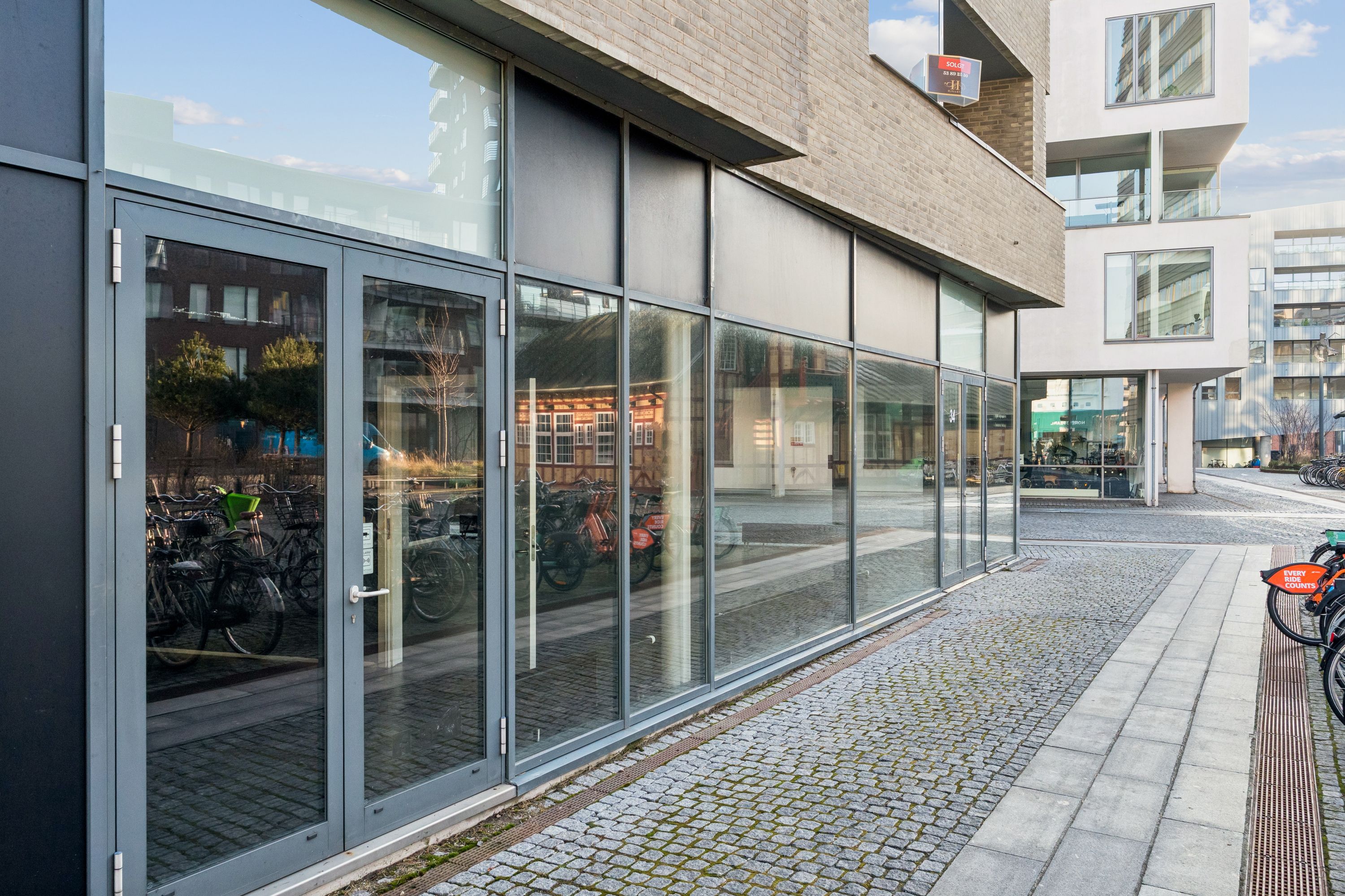 Moderne butik til leje med store vinduer og indgangsdøre, der reflekterer bygninger og cykler på Amerika Plads i København. Solrig dag med lyse himmel og glasvægge.