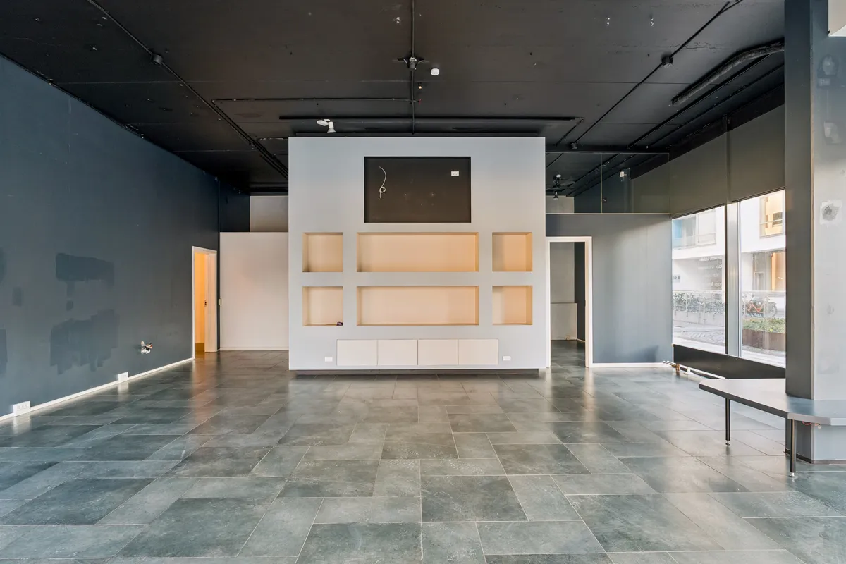 En tom butik med gulv og vægge i grå og hvid, et stort glasvindue og en række hylder i væggen, der vender mod vejen