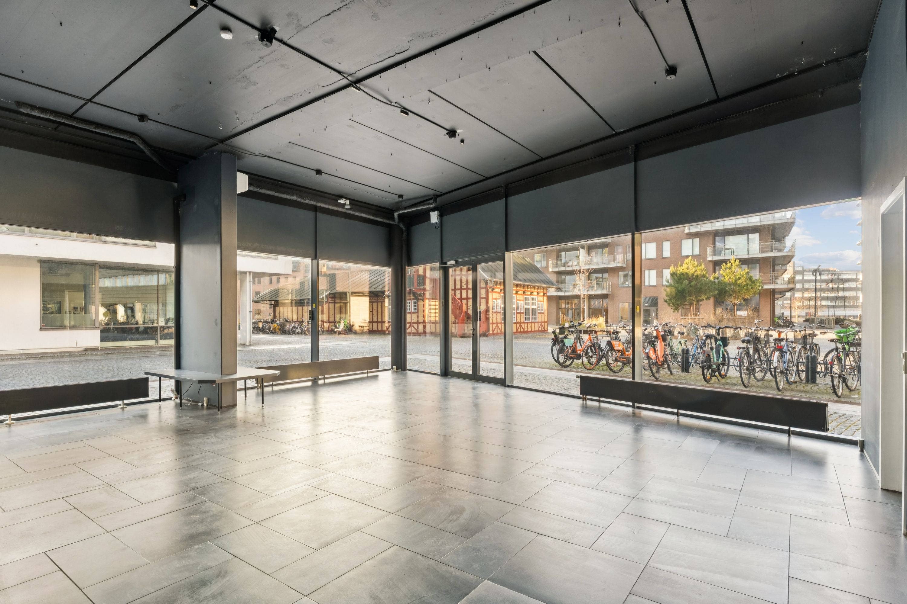 Rummelig showroom med store vinduer og trægulv. To stole og to bænke står i udstyret. Store glasdøre og vinduer giver masser af lys og udsigt til gaden med parkerede cykler