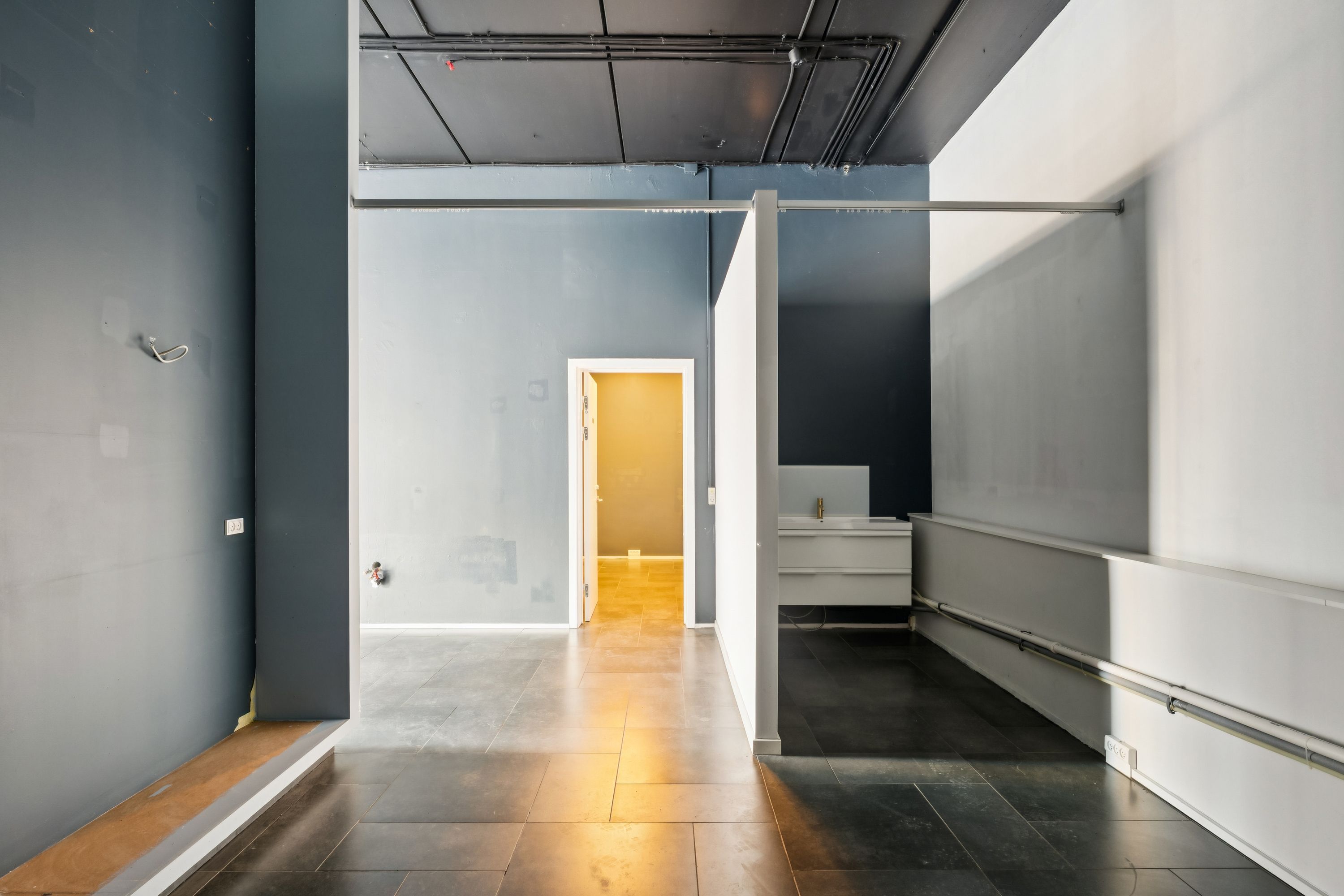 En stor åben rummelig gang med hvid og grå vægge og et åbent gulv med lys og skygge fra døråbningen