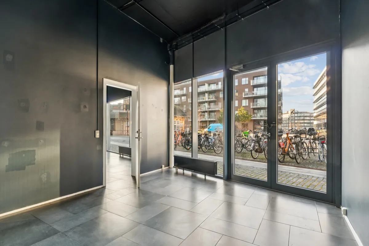 Rummelige lokaler til leje i København med store vinduer og udsigt til gaden og parkerede cykler. Indgangsdøren er åben, og der er flere cykler parkeret udenfor.