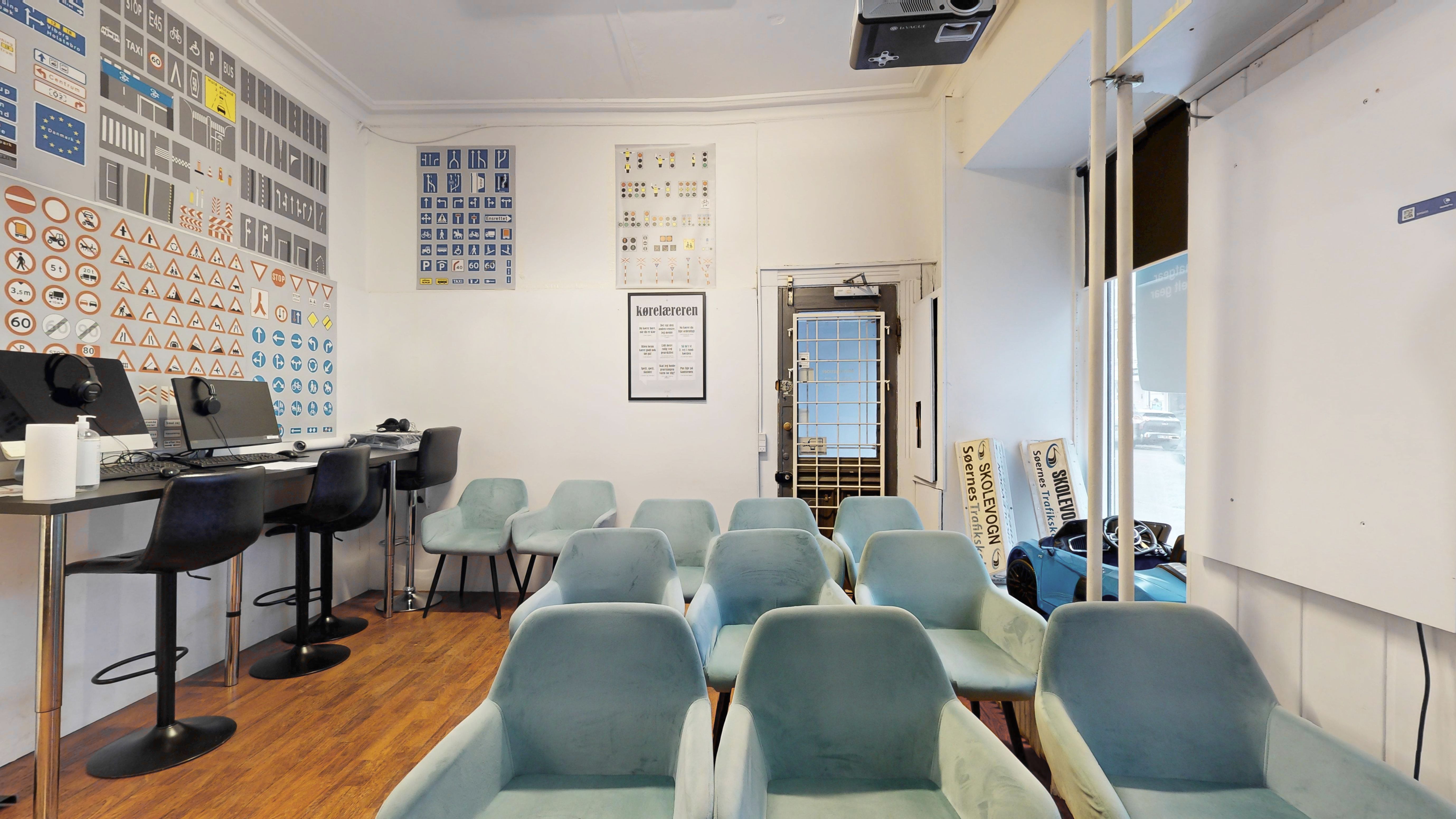 Et kontorlokale med lyse møbler, en monitor og et projektorloft. Der er plakater med trafikskilte og regler på væggen. Der er også en dør, der fører til udendørsområdet med en bil og en annonceplakat