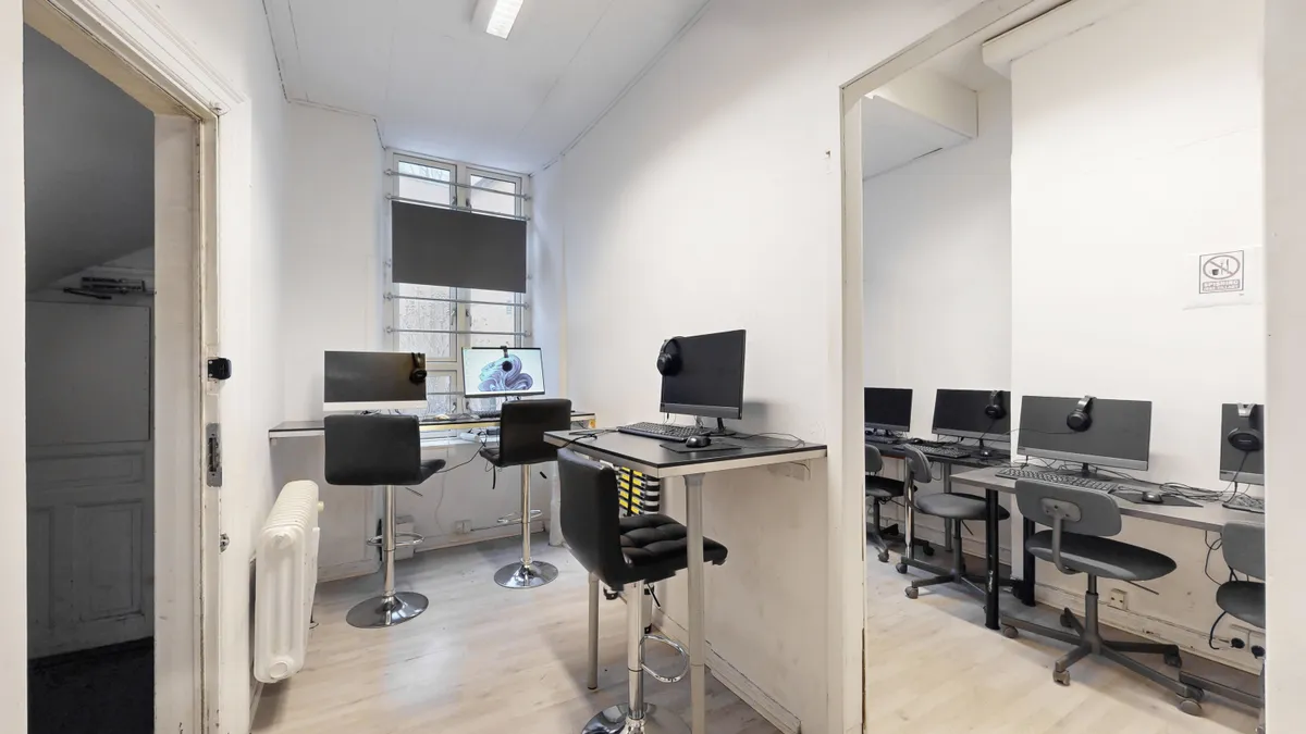 Kontorrum med to borde og flere skriveborde og stole med skærme og computere og en radiator