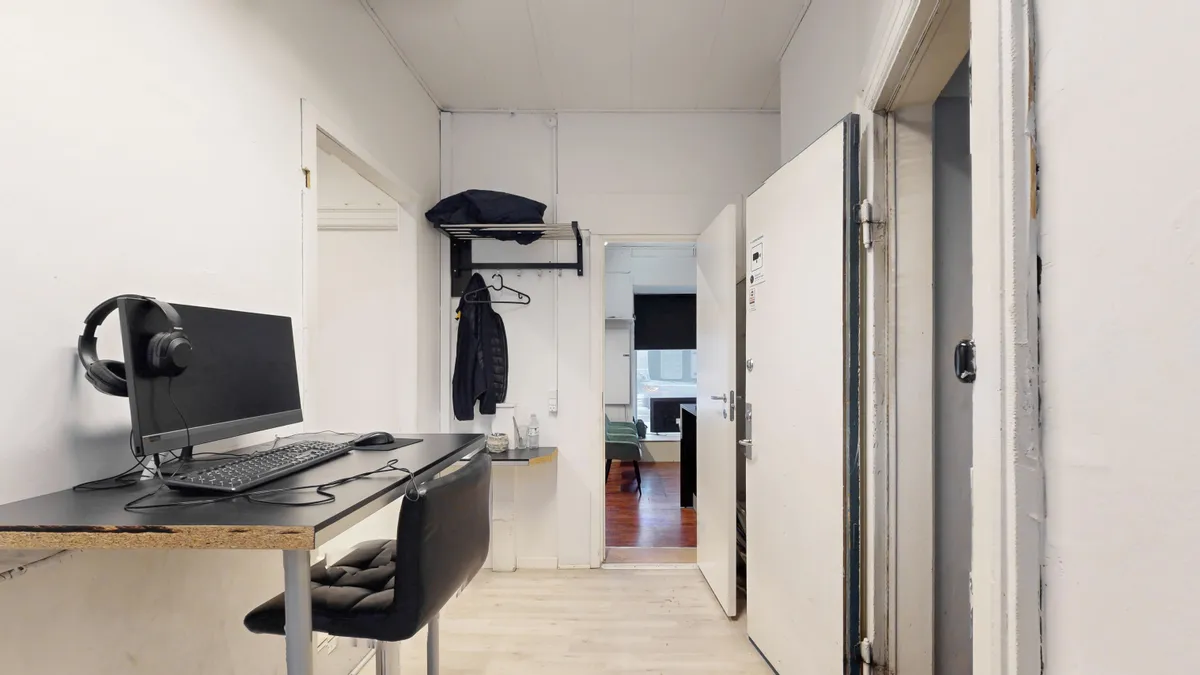 Kontorrum med skrivebord, computer, headset og stol og tøjhænge i hjørnet