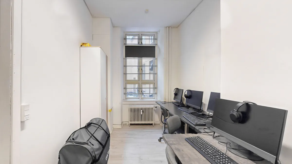 Et kontorlokale med tre computere og deres tilhørende skærme, tastature og mus samt hovedtelefoner placeret på et bord