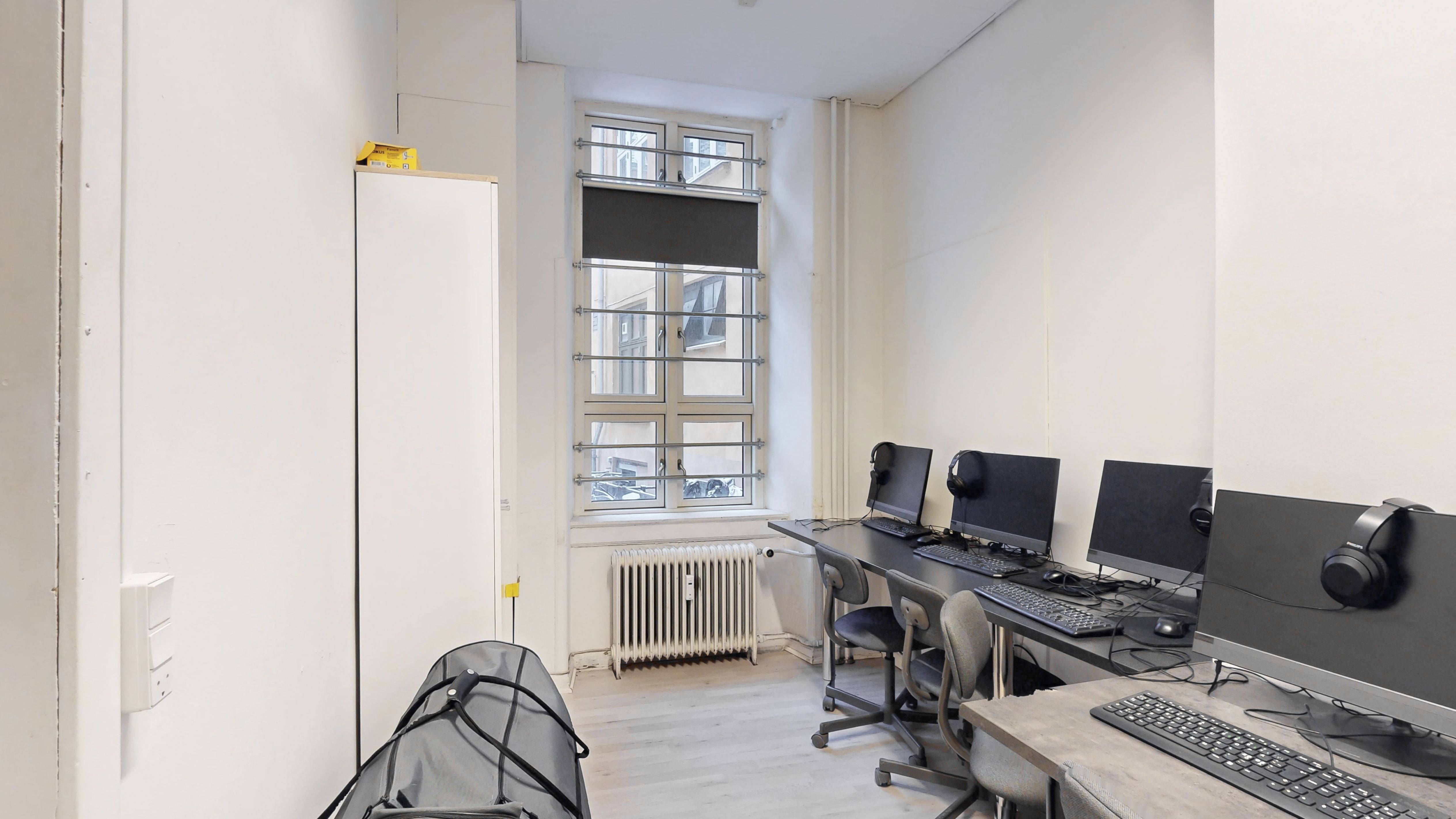 Kontorrum med to borde, hver med to skriveborde, computere, mus, tastaturer, og høretelefoner. Et skab står mod væggen, og en radiator er installeret på gulvet.