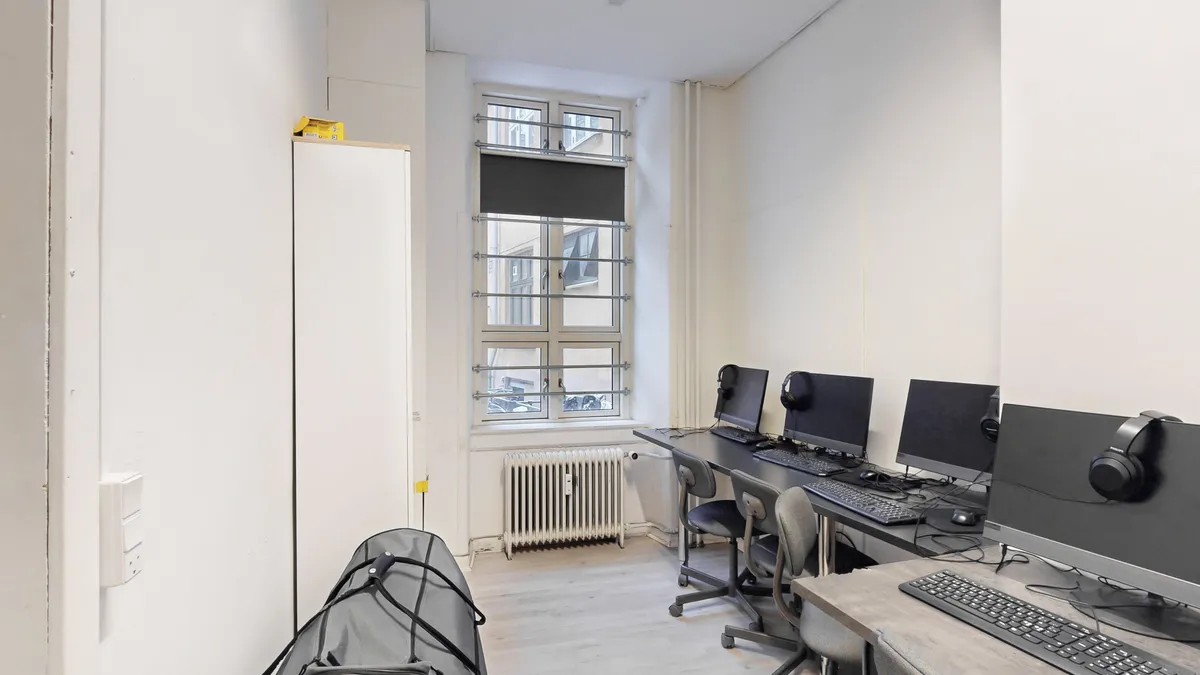 Kontorrum med to borde, hver med to skriveborde, computere, mus, tastaturer, og høretelefoner. Et skab står mod væggen, og en radiator er installeret på gulvet.