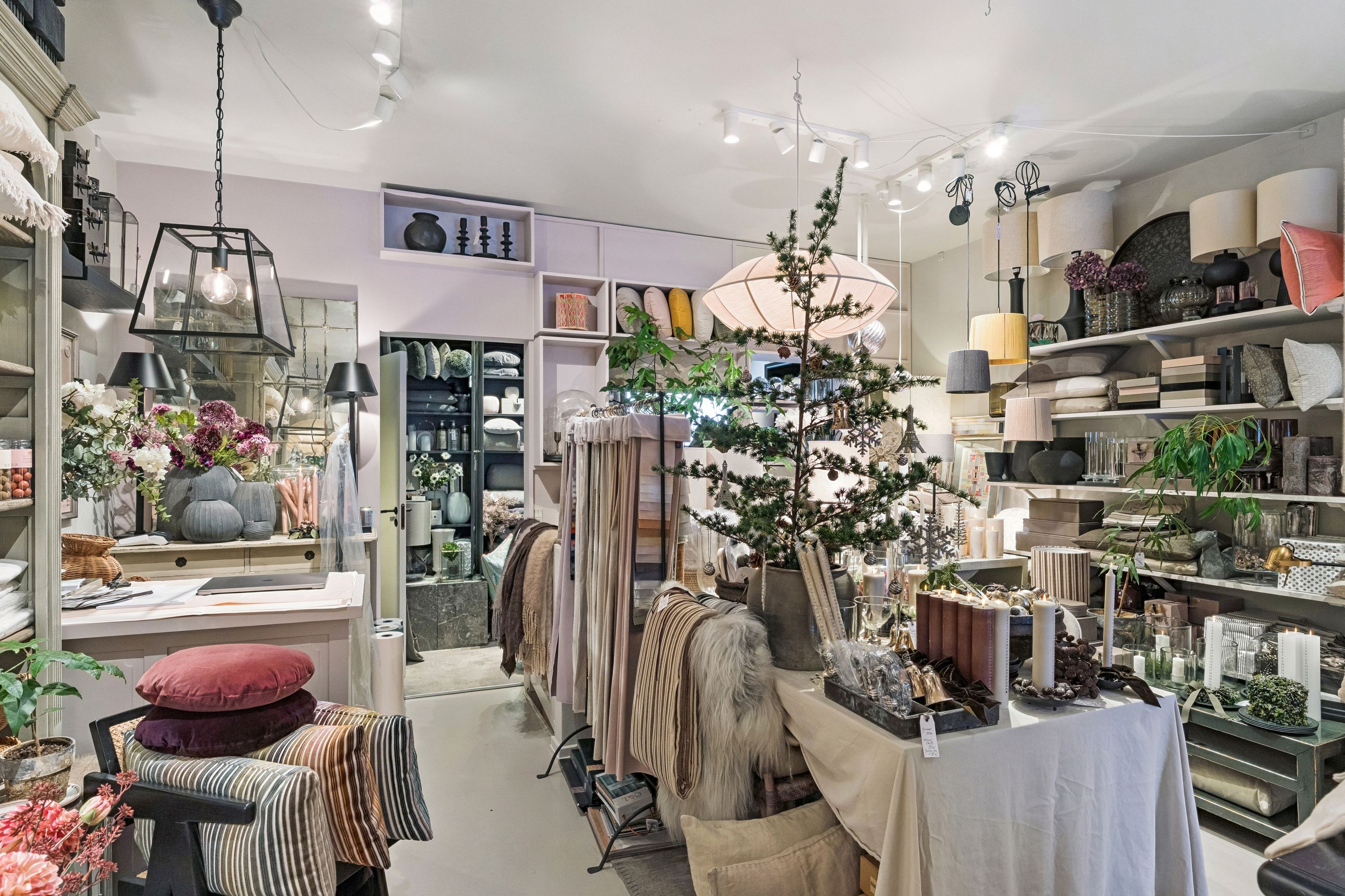 Indvendørs butik med hyggeligt møbelsortiment, lamper, puder og blomster. Møbler og tekstiler er udstillet på hylder og stole. Puder er blevet lagt på stole og borddækket er dækket med en klud