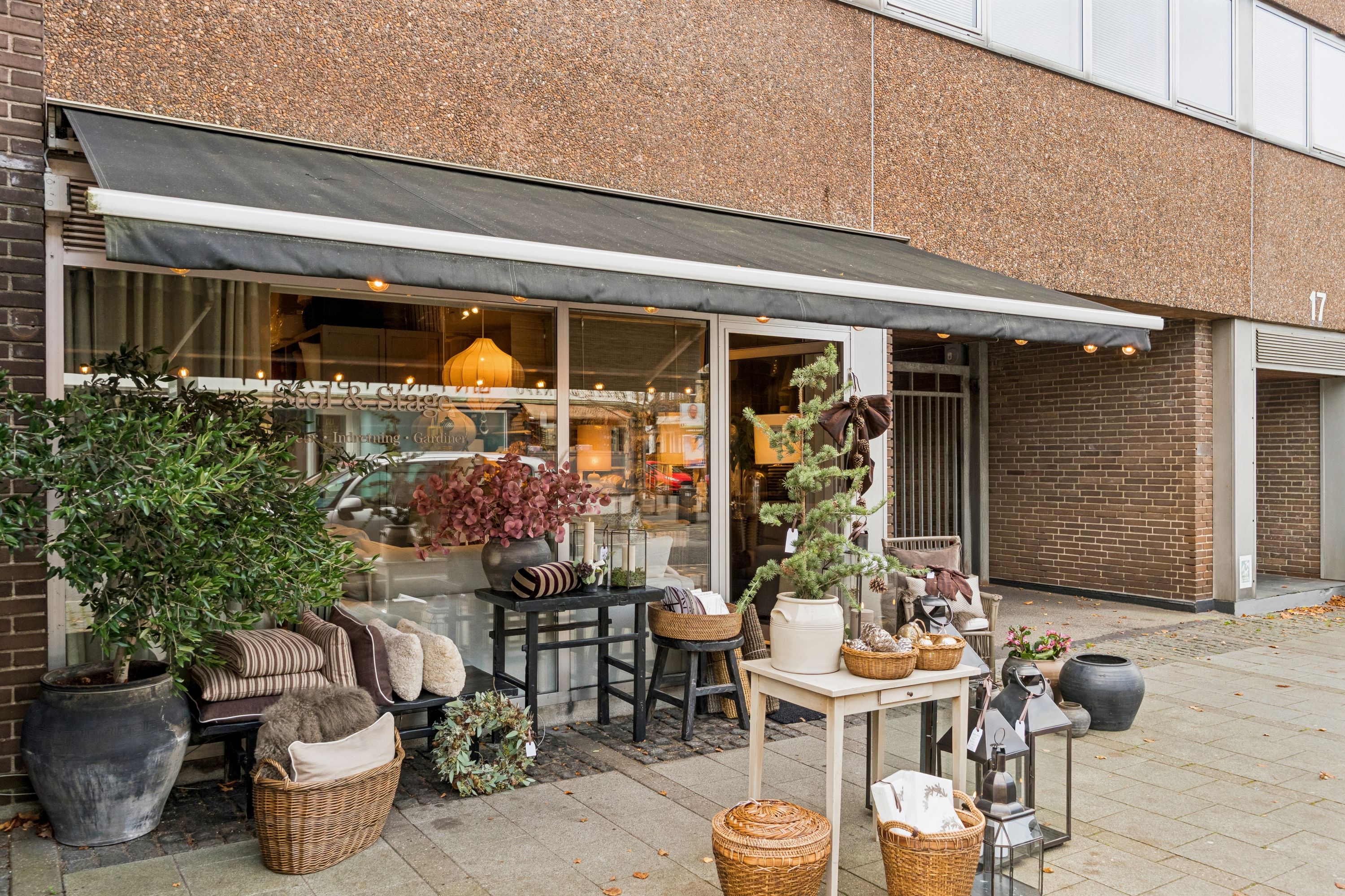 Et moderne butikslokal med glasvinduer, en terrasse med møbler og plantepoter og et sort audrab på en terrasse i forgrunden.