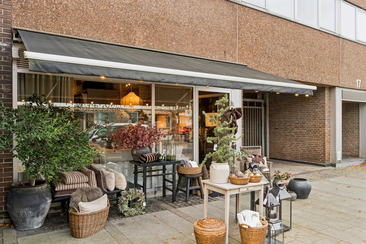 Et moderne butikslokal med glasvinduer, en terrasse med møbler og plantepoter og et sort audrab på en terrasse i forgrunden.