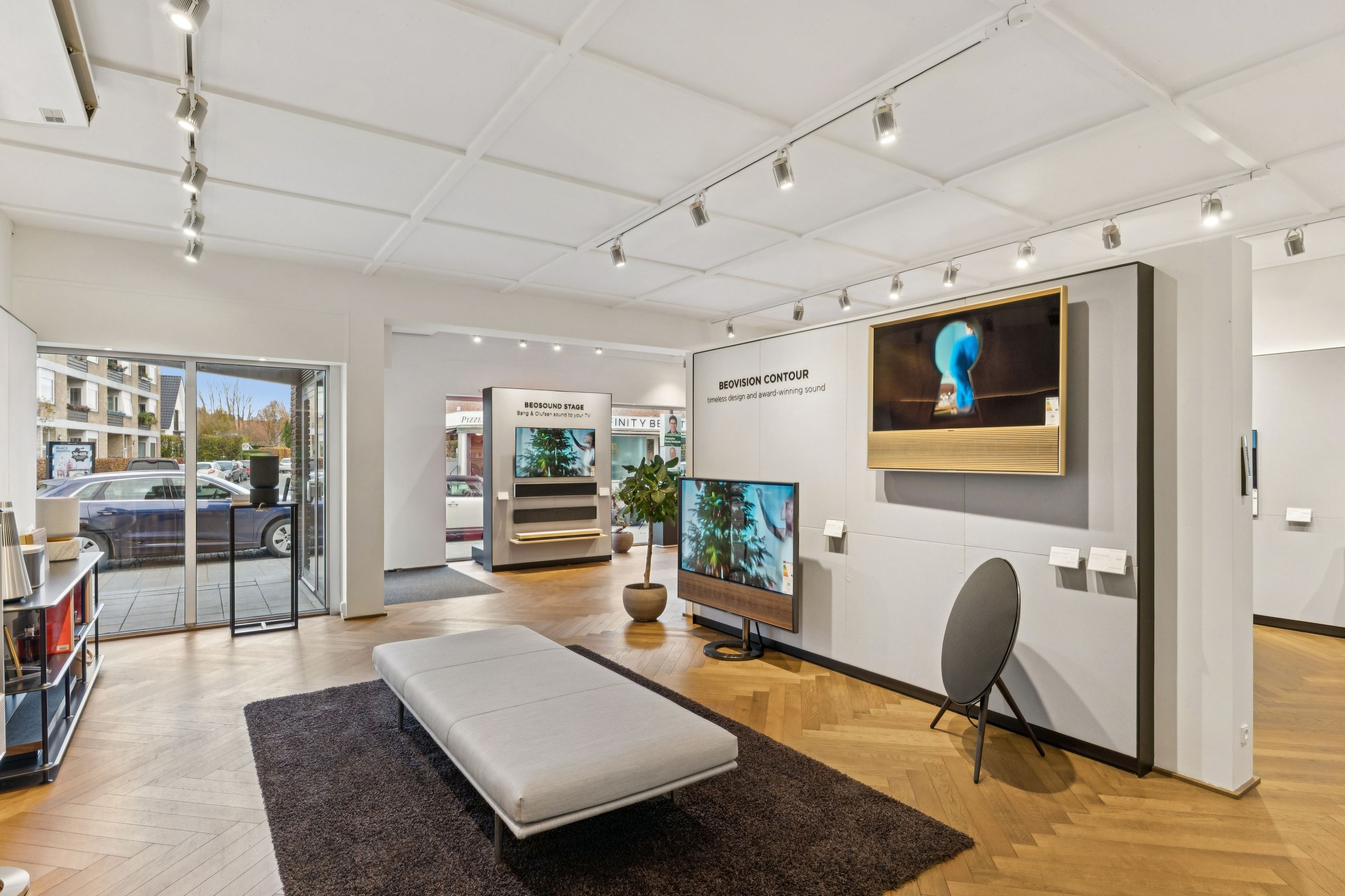 Store vinduer med udsigt til parkeringsplads og biler, tv-skærme og møbler i minimalistisk interiør med trægulv og højt loft