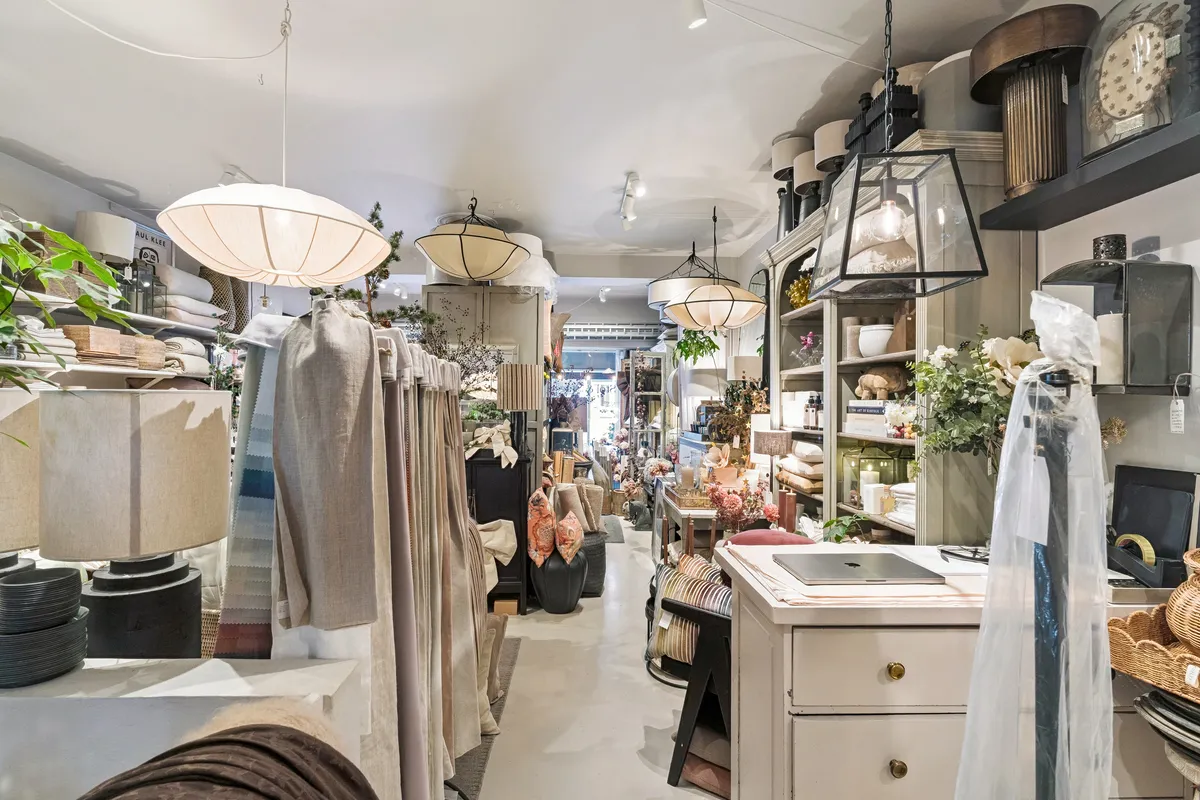 Butik med møbler og tekstiler, hængende lamper, lysekroner og hylder med varer og blomster i en smal gang mellem høje hylder og smukke lysekroner