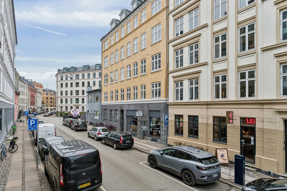 En bilparkeret gade i København med parkerede biler og cykler. Bygningerne med høj opholdsværdi og moderne indretning. En bil er i gang med at lade ved en elektrisk ladestøtte.