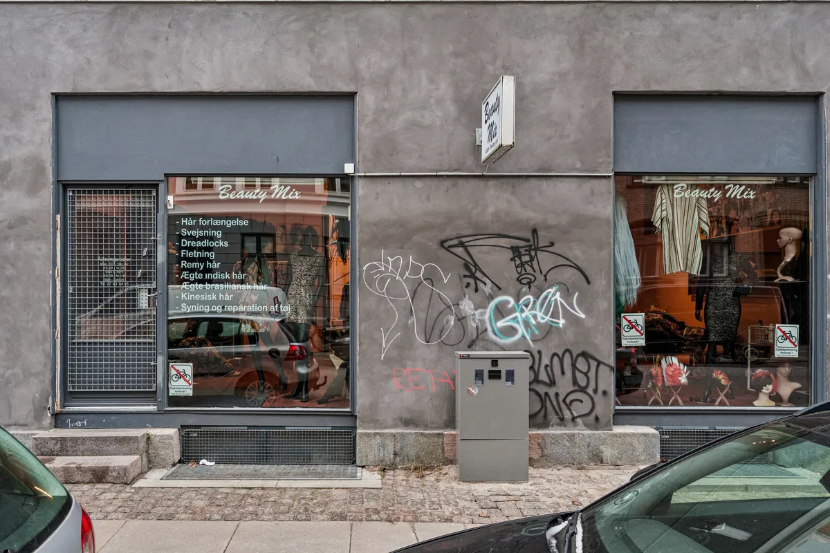 Butik med Beauty Mix-skilt og forskellige tjenester som hårforlængelse og drædlocks. Butikken har graffiti på væggen og nogle reklamer i vinduet.