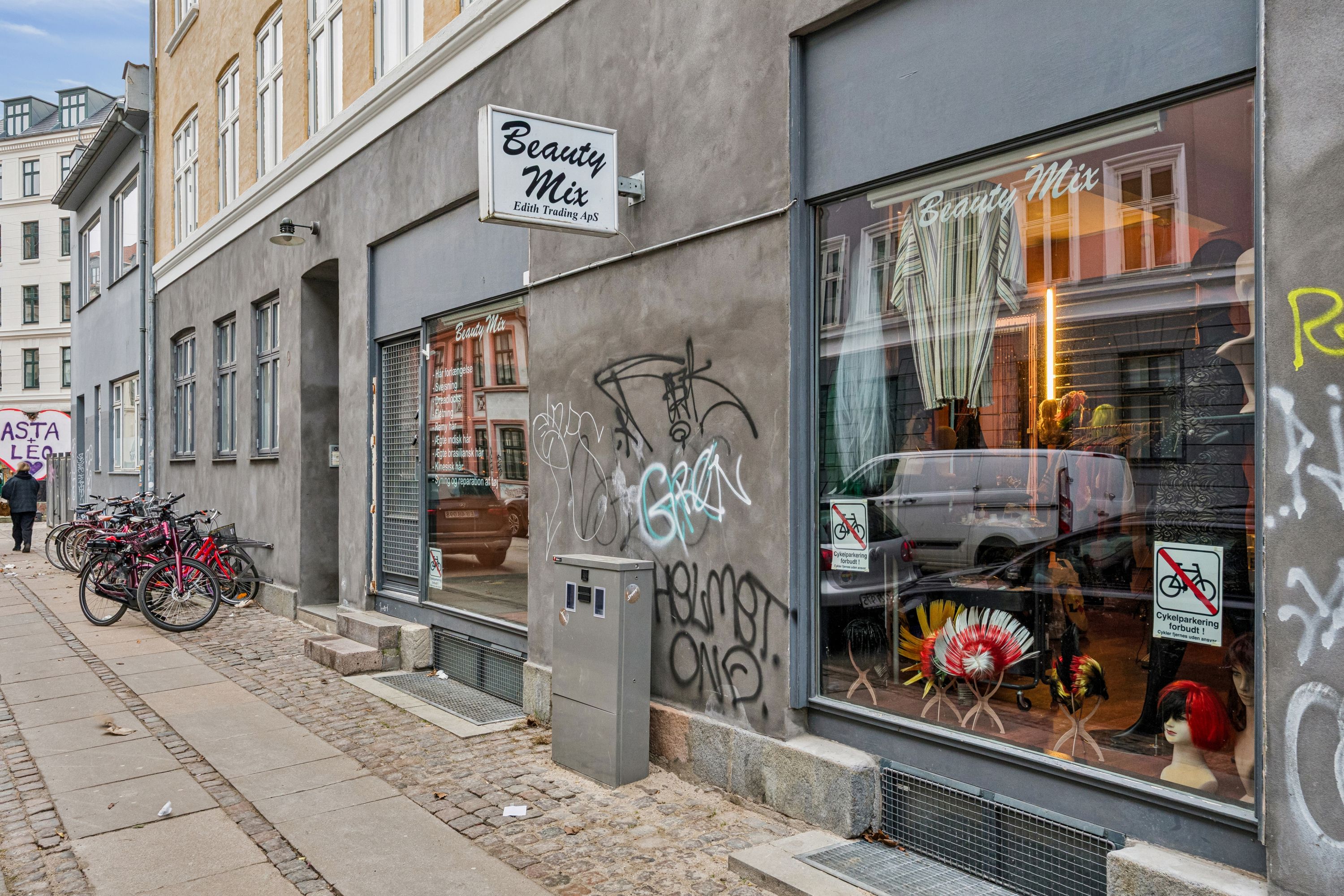 Butik til salg på Viktoriagade 9 i København V. Skilte med navnet Beauty Mix og graffiti på facaden. Cykler parkeret udenfor og vinduer med refleksioner af biler og folk
