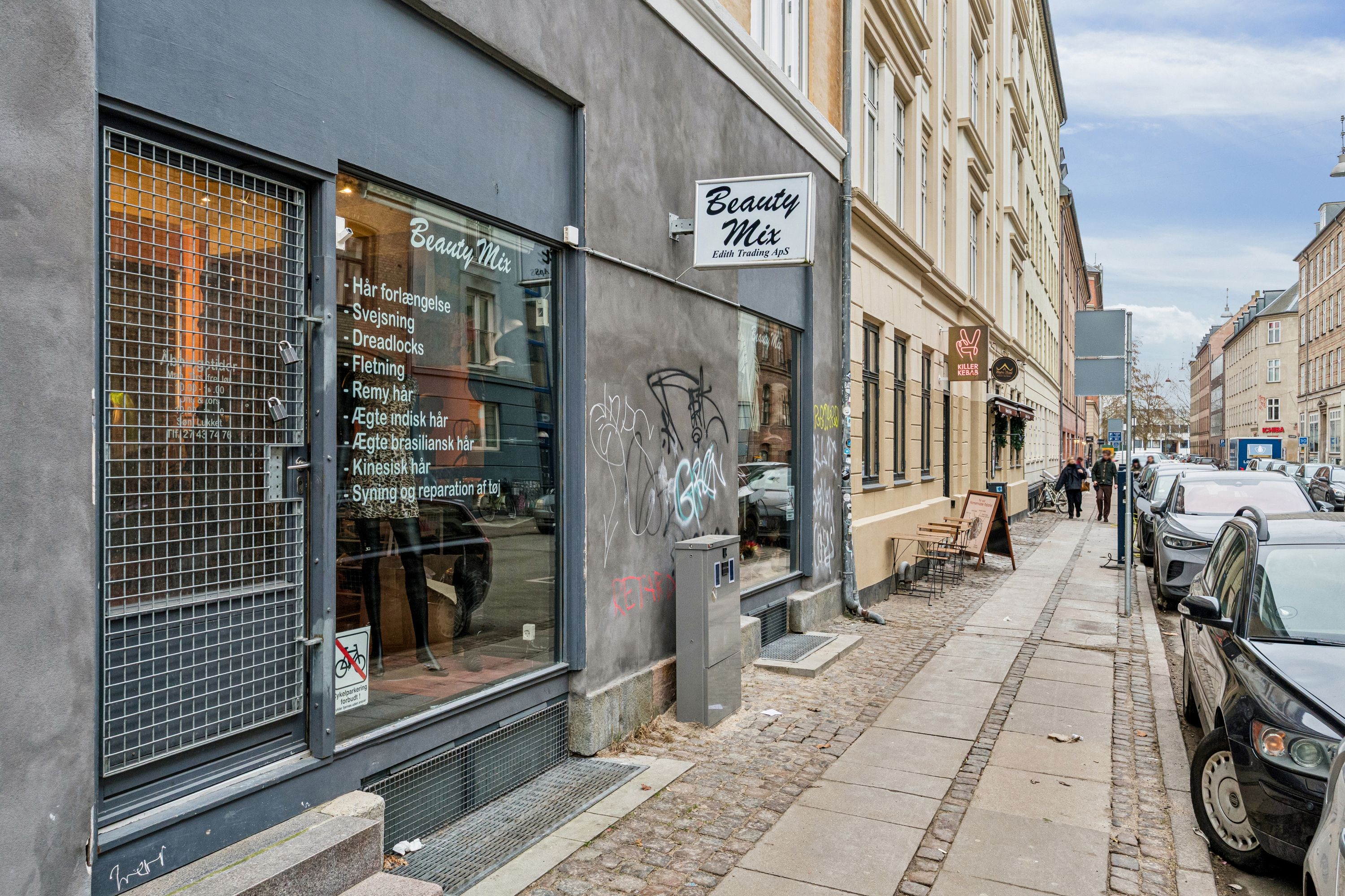 En Beauty Mix butik med et skilt over indgangen og en række tjenester som svejsning og kinesisk. En række biler er parkeret på siden af gågaden.