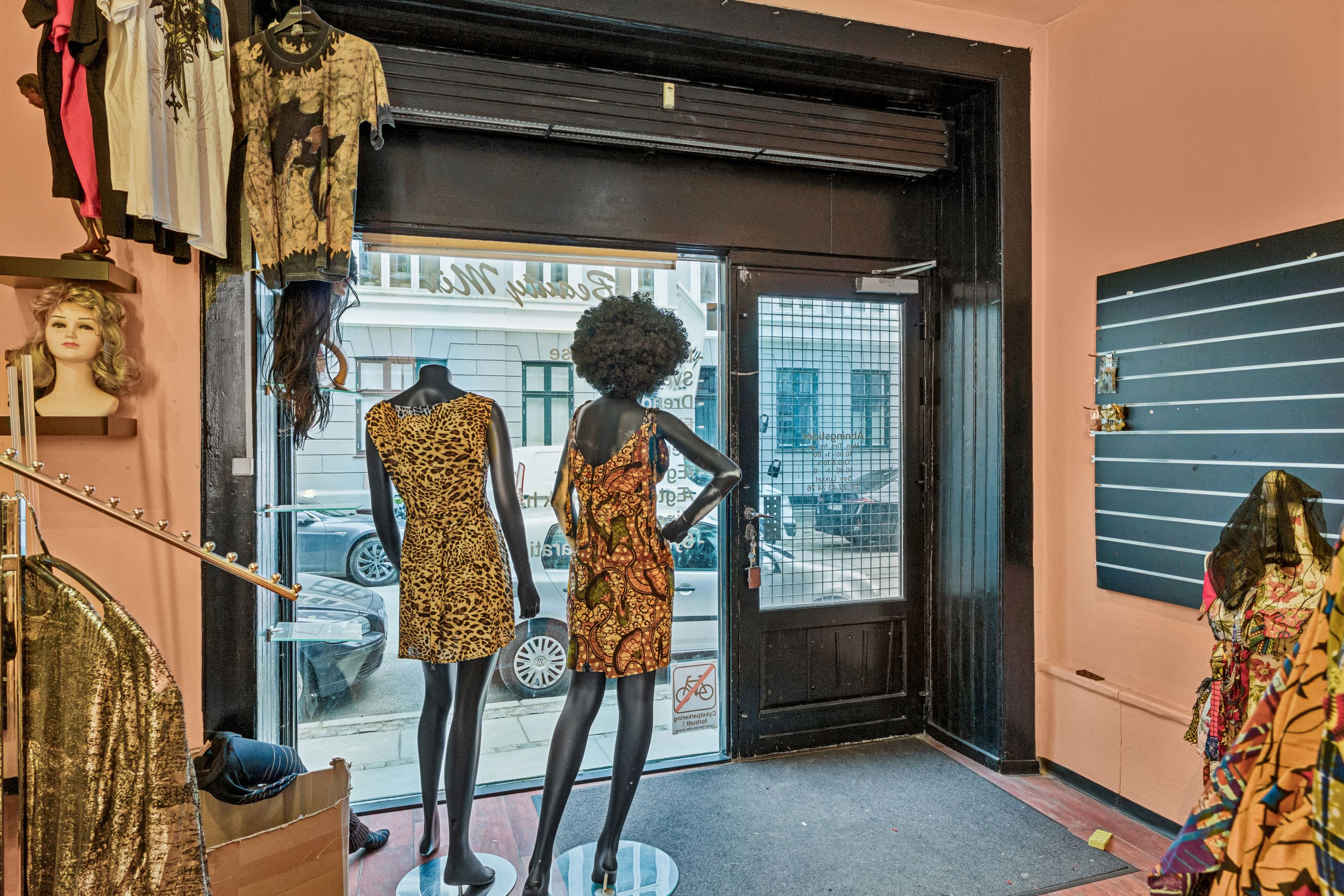 En butik med en åben dør viser to sort tøjdukker i farverige kjoler og tøj fra loftet og et spisestuebord med møbler og en spisestue med et tøjstativ