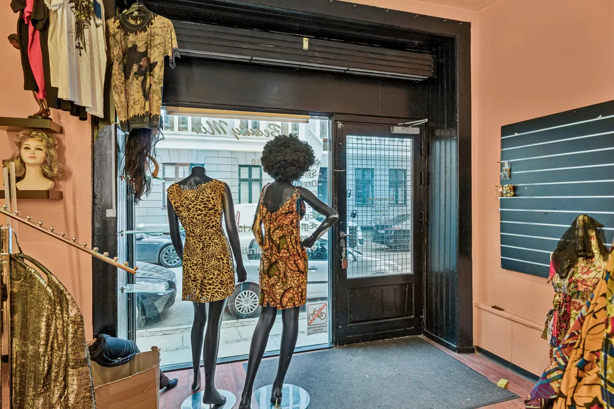 En butik med en åben dør viser to sort tøjdukker i farverige kjoler og tøj fra loftet og et spisestuebord med møbler og en spisestue med et tøjstativ