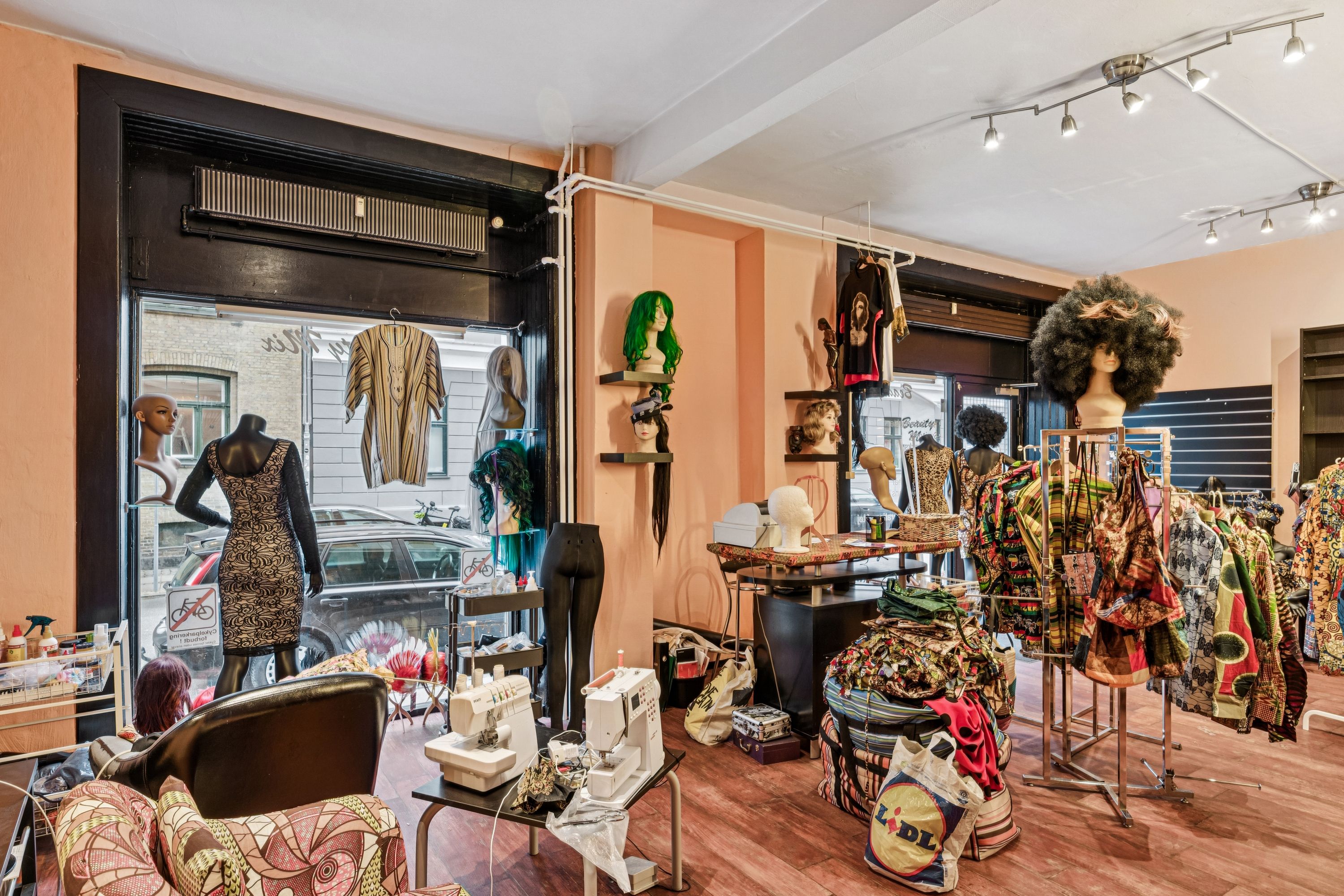 Københavnsk butik med mange tøjstykker, mønstre og manequins. To symaskiner og stole i forgrunden. Glasskure med biler og bygninger bagved