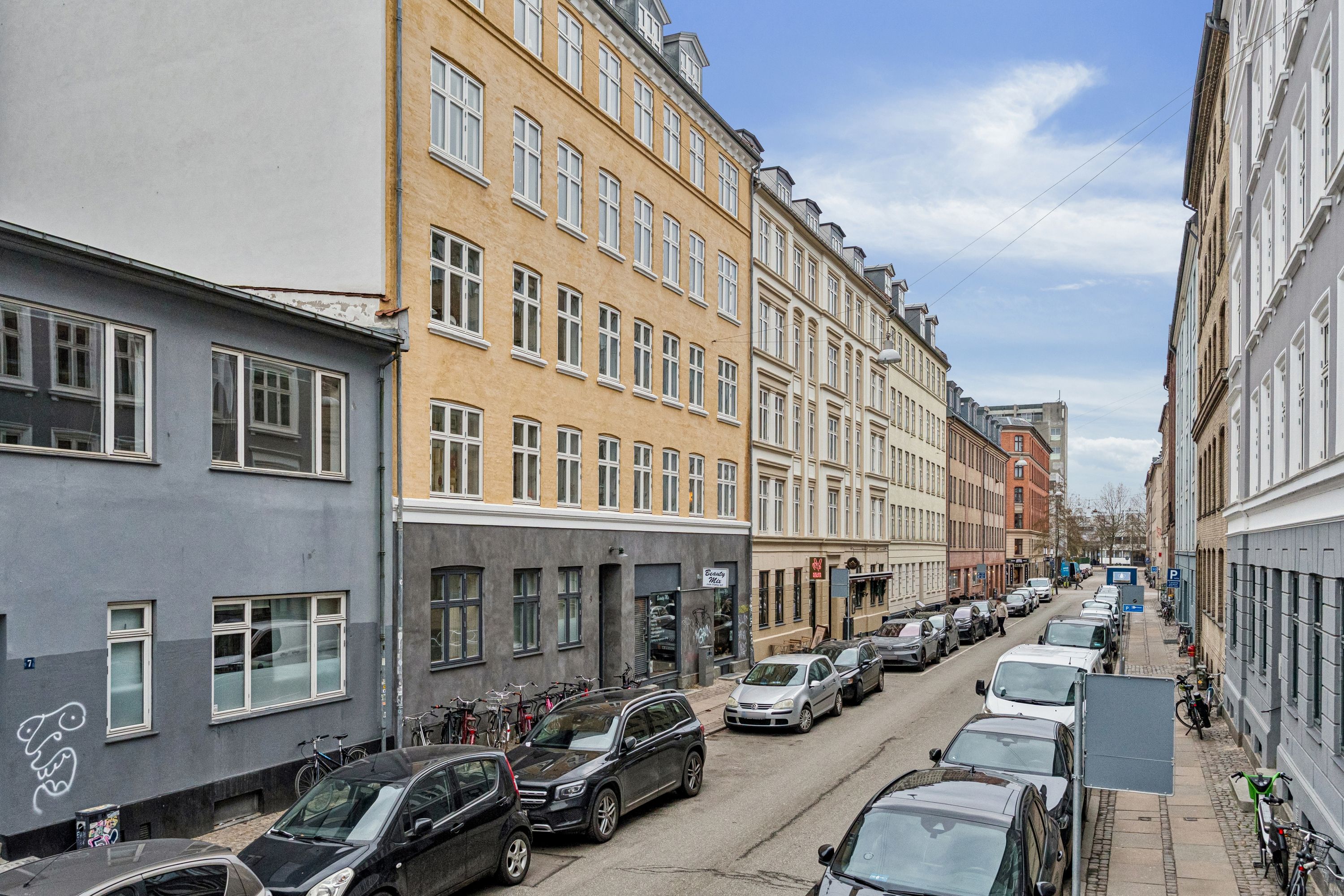 Adgangsvej med parkerede biler og cykler og flere butikslokaler med vinduer og glasdøre, hvor der er graffiti på en af facaderne