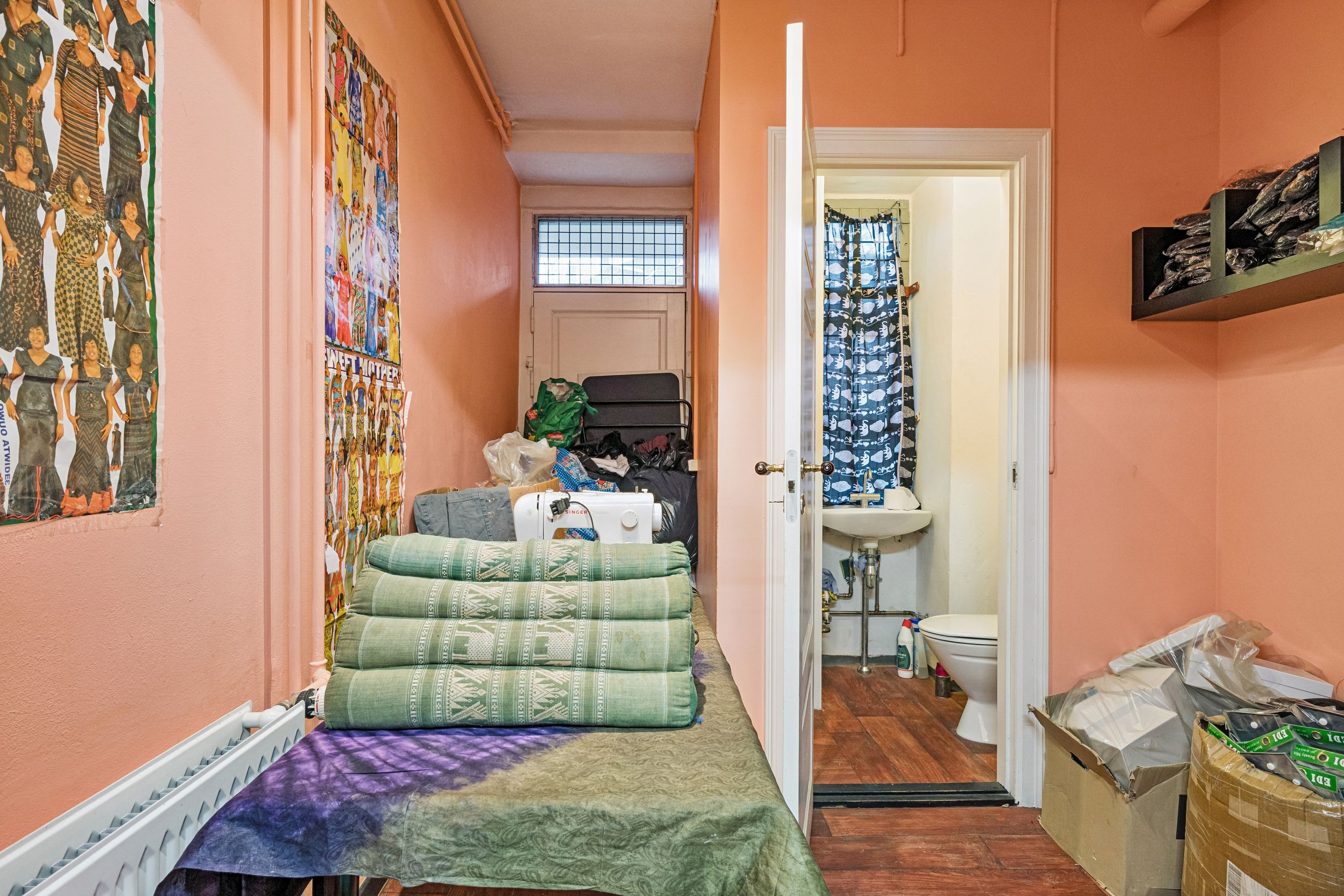 En række madrasser og tøj på en seng i en stue med lyserøde vægge og en åben dør til et badeværelse med toilet, skænk og vinduesgardin