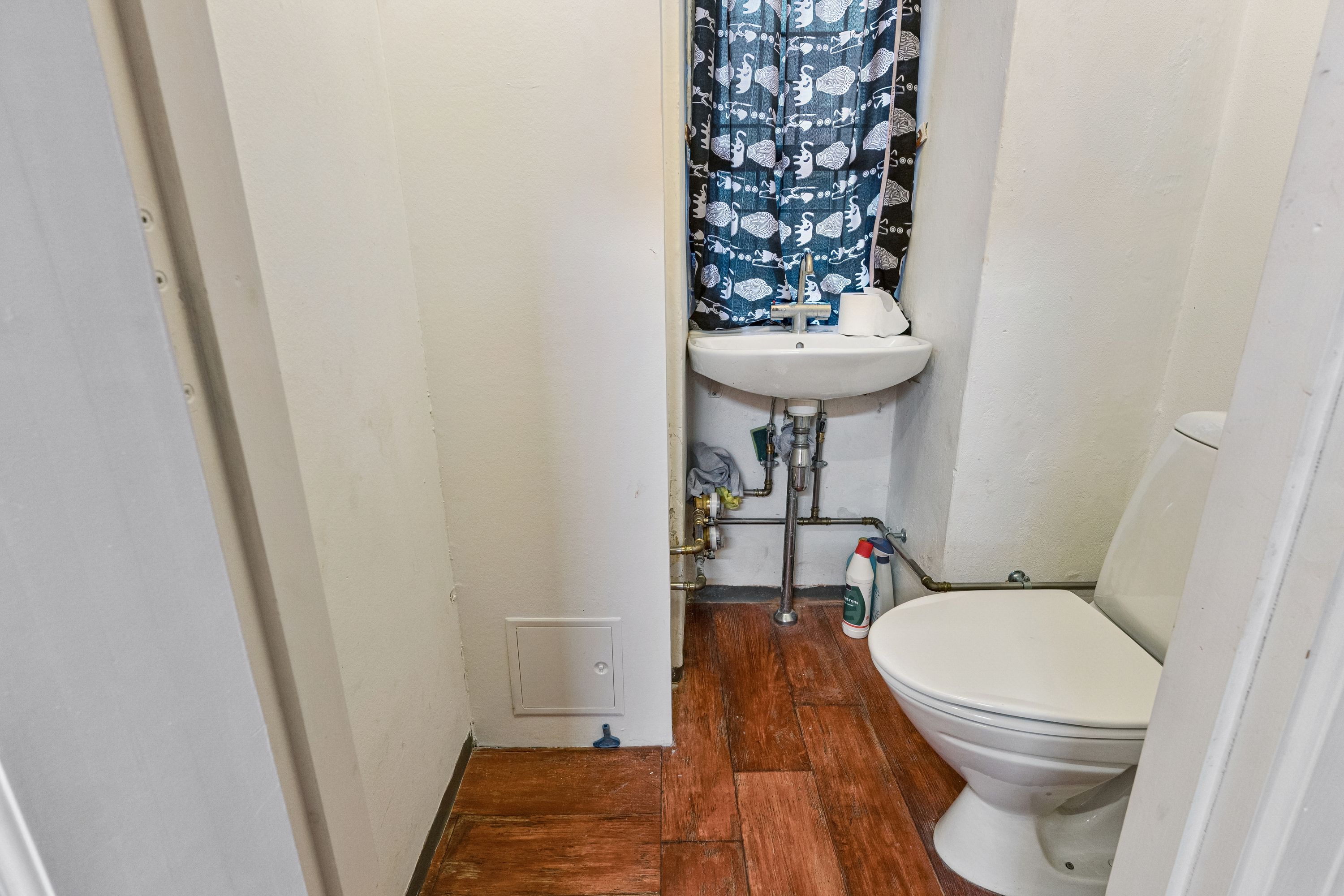 Et lille badeværelse med toilet, hvidt badekar, en hvid håndvask med blå kurtiser og to rengøringsmidler på gulvet. Gulvet er lavet af træ.