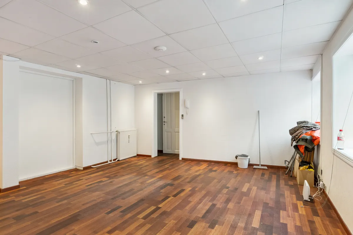 En tom, hvidvægget kontorrum med trægulv og vinduesvisning. Rummet er tomt med en bøjle og en kuffert ved siden af vinduet. Der er også en stang med tøj og en flaske på vindueskarme.