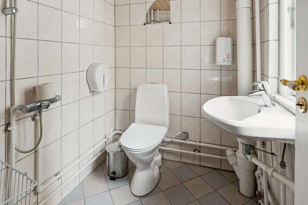 En moderne, pænt ren toiletrum med lyse beige fliser, hvidt toilet og sink samt et lille spejl. Håndklæder og en håndklædetørrer er placeret ved siden af.