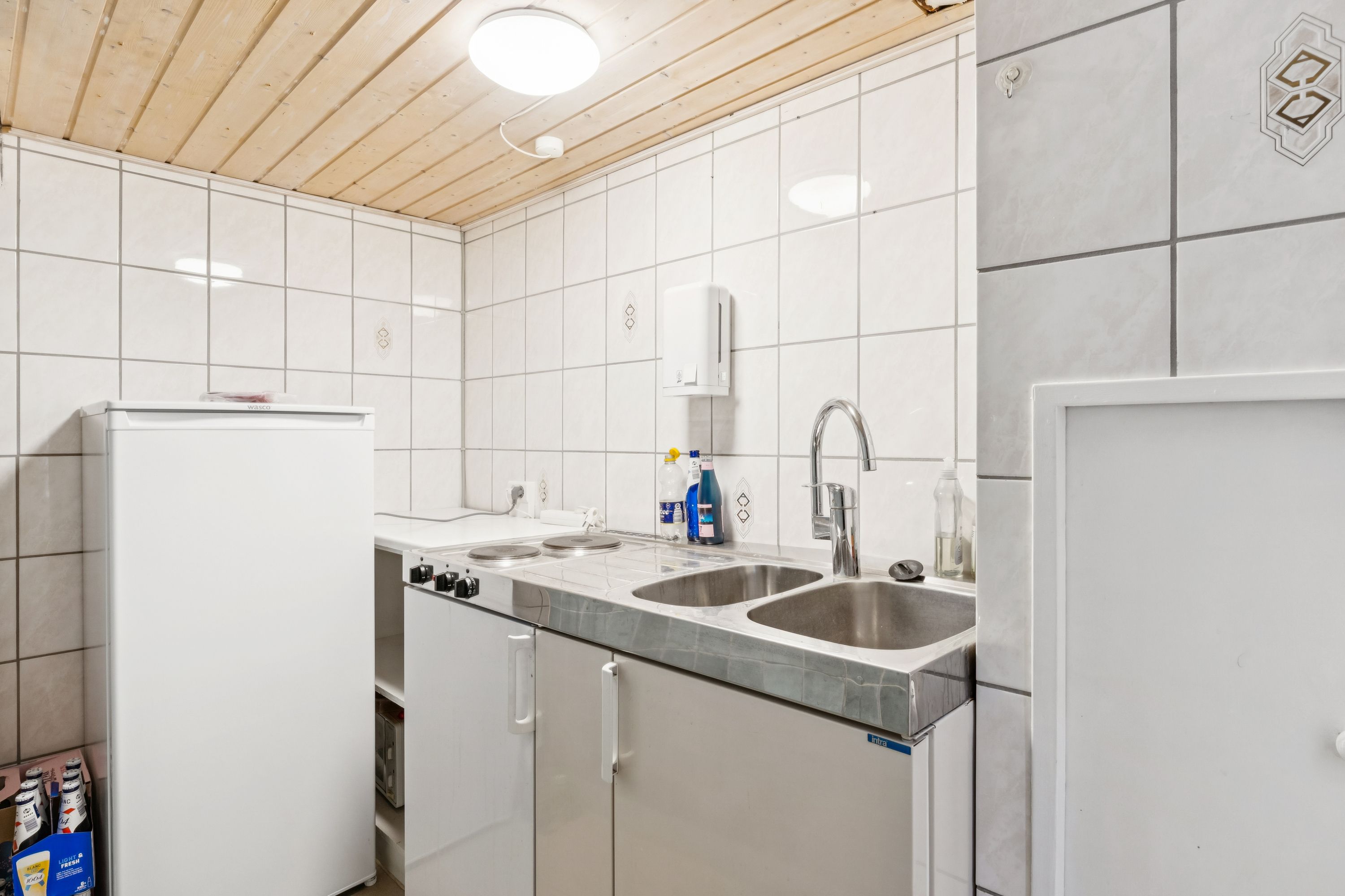 Alt-tekst: En rent holdt køkken med hvide vægge og loft, en indbygget komfur, en hvid køleskab og to lyse, runde, særegne lysekroner.