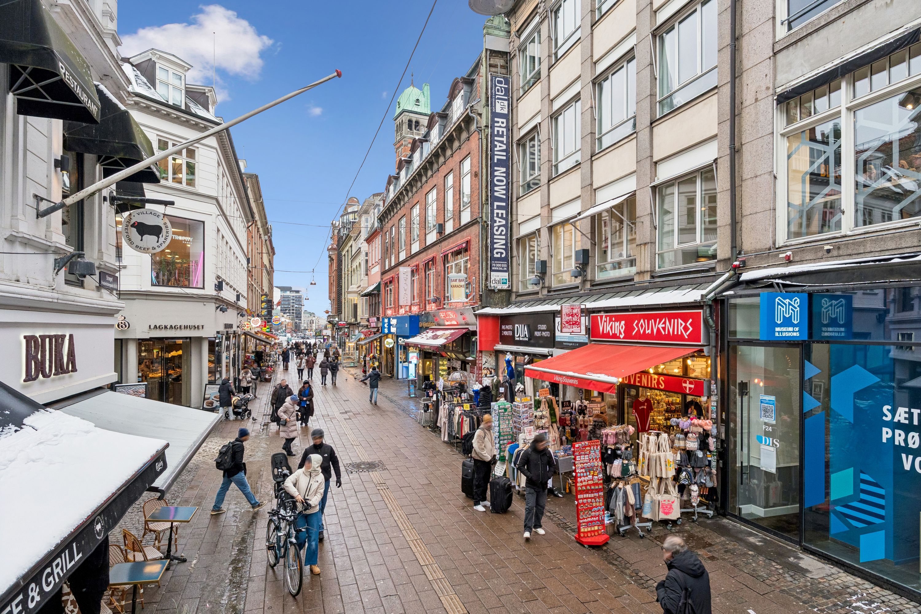 Folk går på en gågade med butikker og kaféer, der er udsmykket med skilte, røde markiser og udstille produkter og gaver