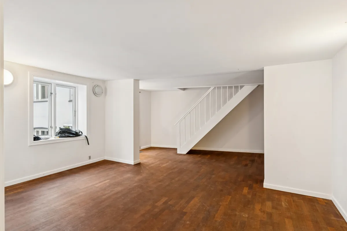 Her ses et tomt kontor med trægulv, hvidt loft og hvide vægge. Stiger til loftsetagen og et enkelt vindue er åbent med en rygsæk på vindueskarmen.