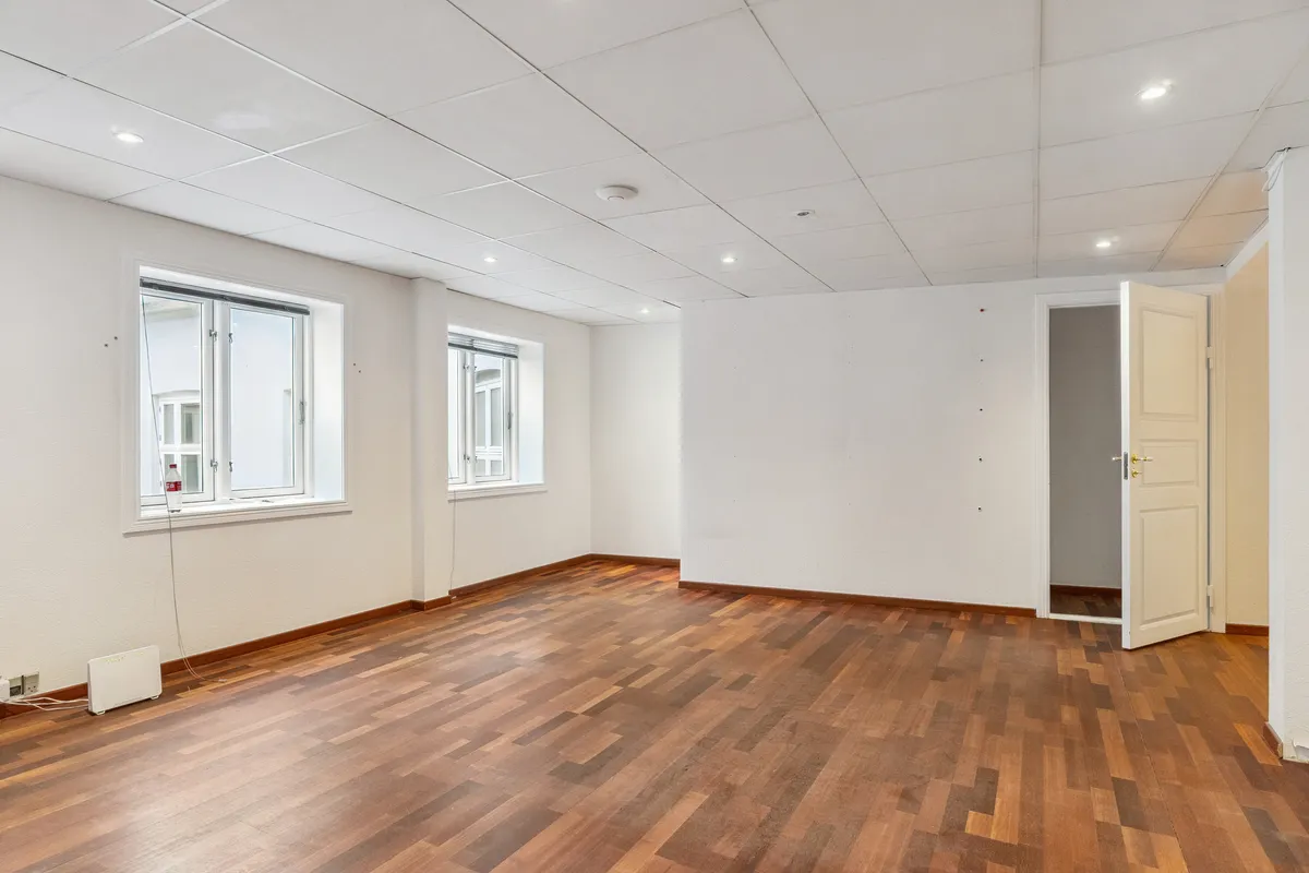 På dette billede ses et tomt kontor med lysegulv, hvidt loft og hvide vægge. Værelset har to store vinduer og to døre, hvoraf den ene er åben.