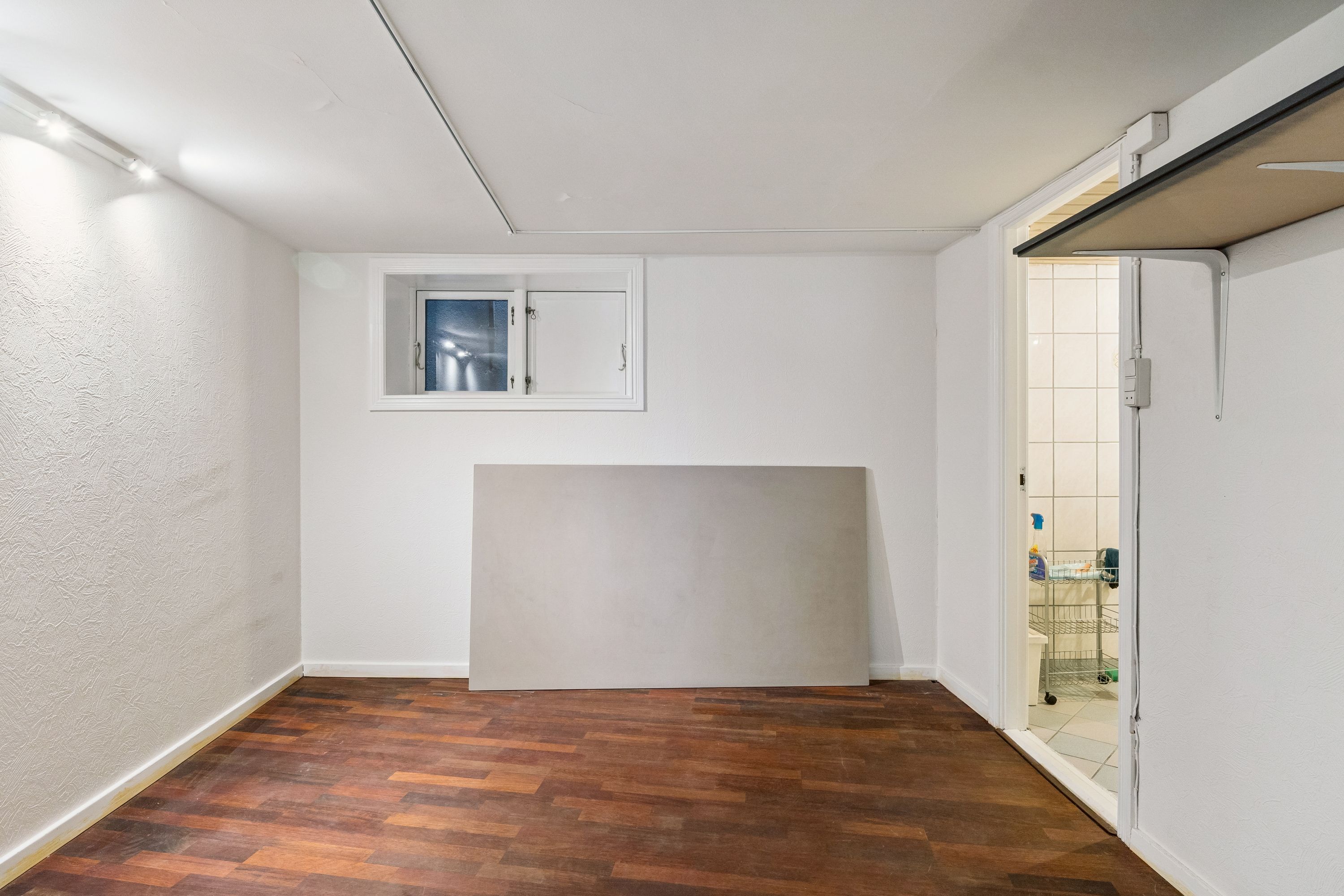 Lejlighedsrum med lyse hvide vægge og gulvtæppe. Rummet er tomt med en hvid væg og et vindue i bagvæggen. Værelset har en åben dør og en metalhylde på væggen