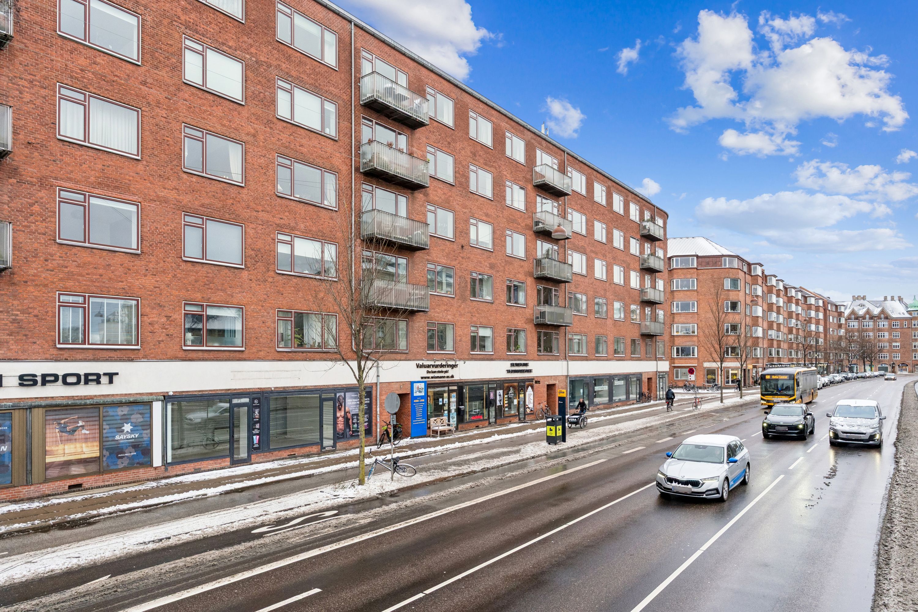 Lejlighed til leje på Åboulevard med sportbutik i første sal, biler kører på vej og sne ligger på stien