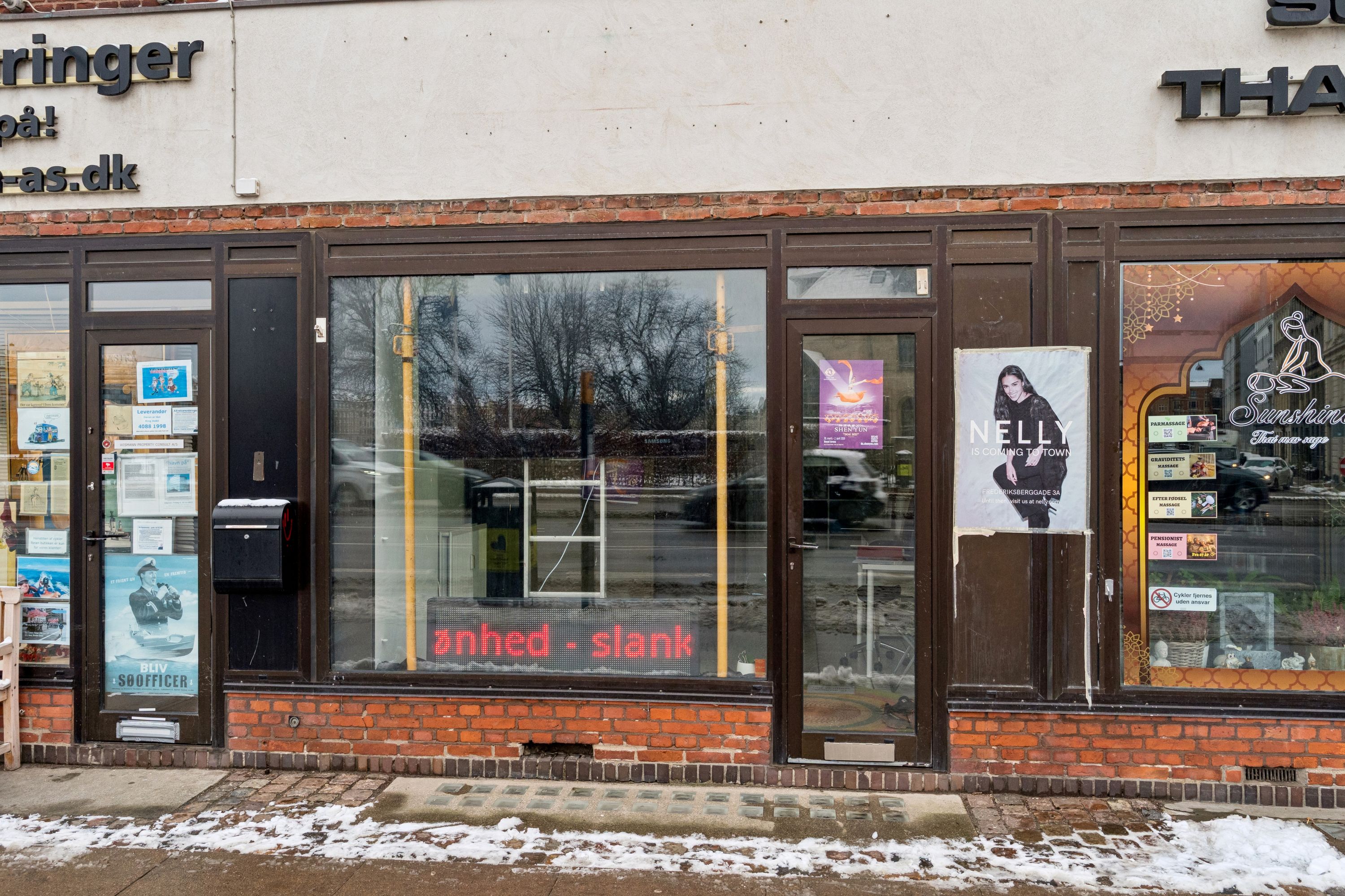 En lille butik med solklat og skilt med teksten Nelly. Butikken er i et hjørne og har røde mursten. Butikken er til leje og har skilt med neonlys og reklameposter i vinduet.