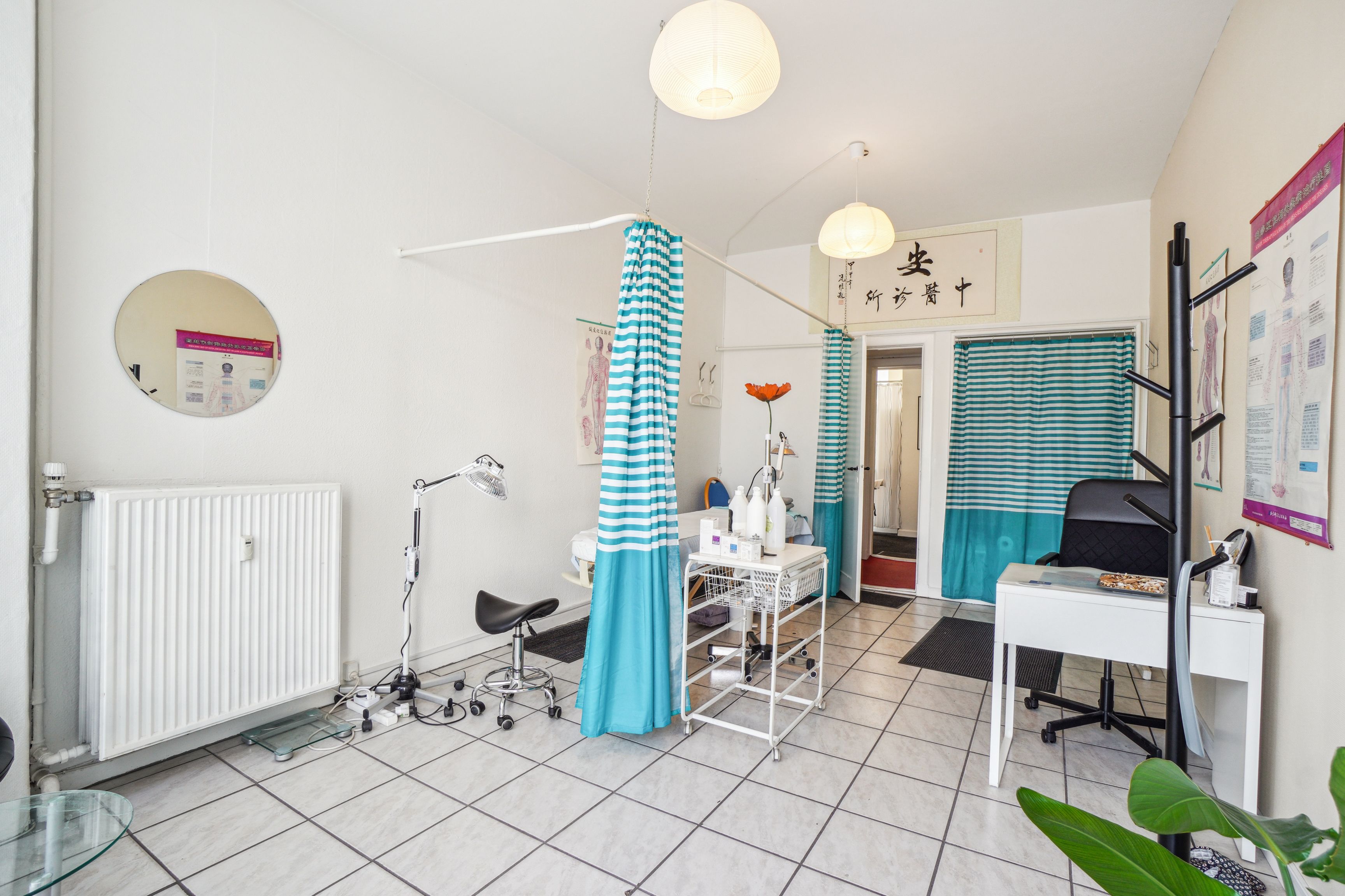 Fin lille butik til leje i København V. På Åboulevard er der et hvidt interiør med lysekroner og vinduesgardiner. Der er et højt gulv med et skråbord og et hvidt bord med et skrivebord.