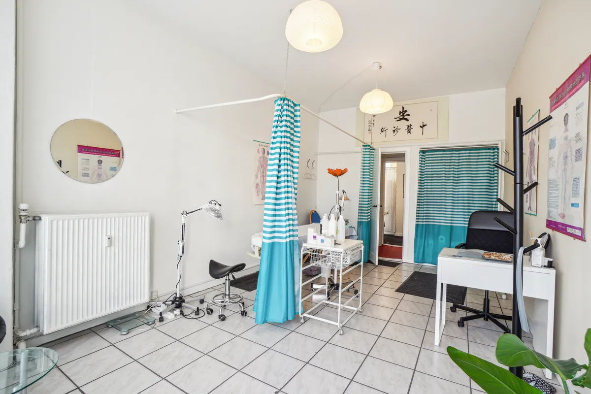 Fin lille butik til leje i København V. På Åboulevard er der et hvidt interiør med lysekroner og vinduesgardiner. Der er et højt gulv med et skråbord og et hvidt bord med et skrivebord.