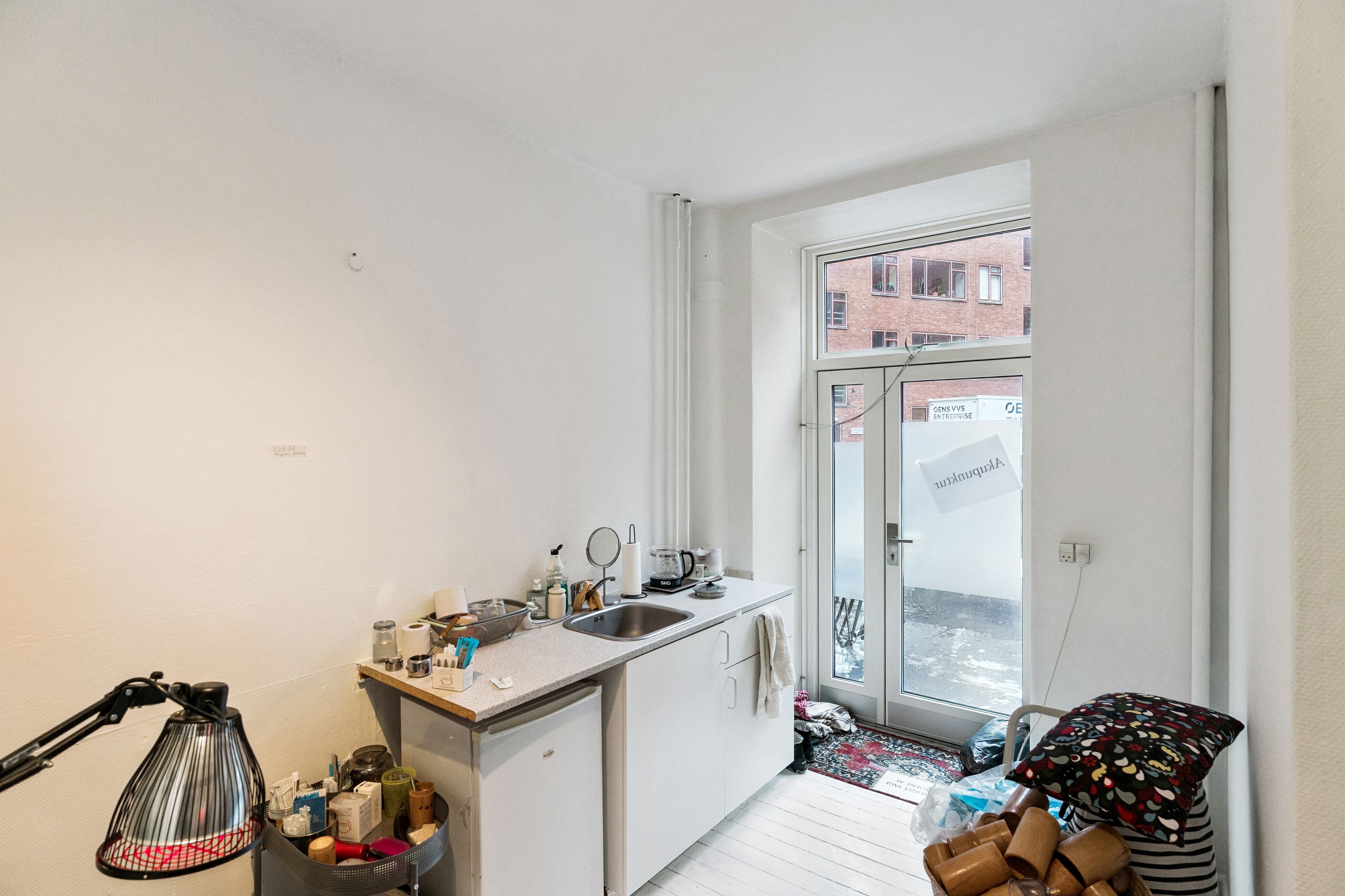 Indeholder lyse og lysegule vægge, hvide køkkenskabe og en dør med skilt, der siger Artisan. Der er en lampe og en kuffert på gulvet.