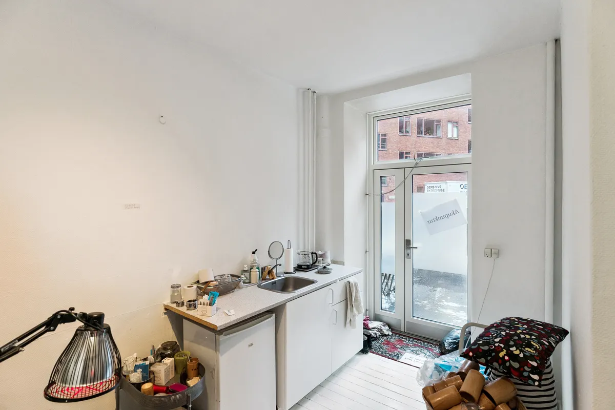 Indeholder lyse og lysegule vægge, hvide køkkenskabe og en dør med skilt, der siger Artisan. Der er en lampe og en kuffert på gulvet.