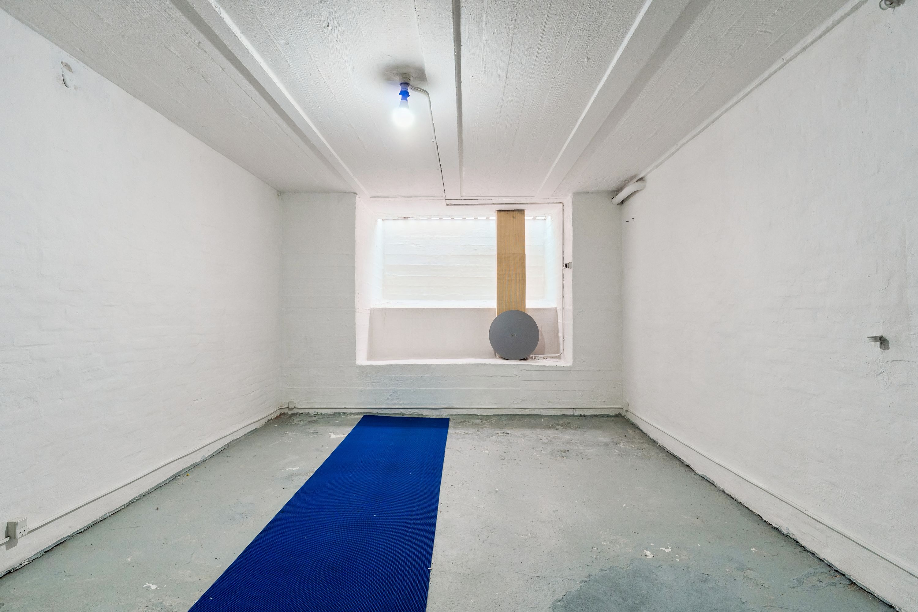 Et tomt rum med hvide vægge og gulv og en simpel indretning med en blå yogamåtte og en rund mørkegrå pude ved et vindue