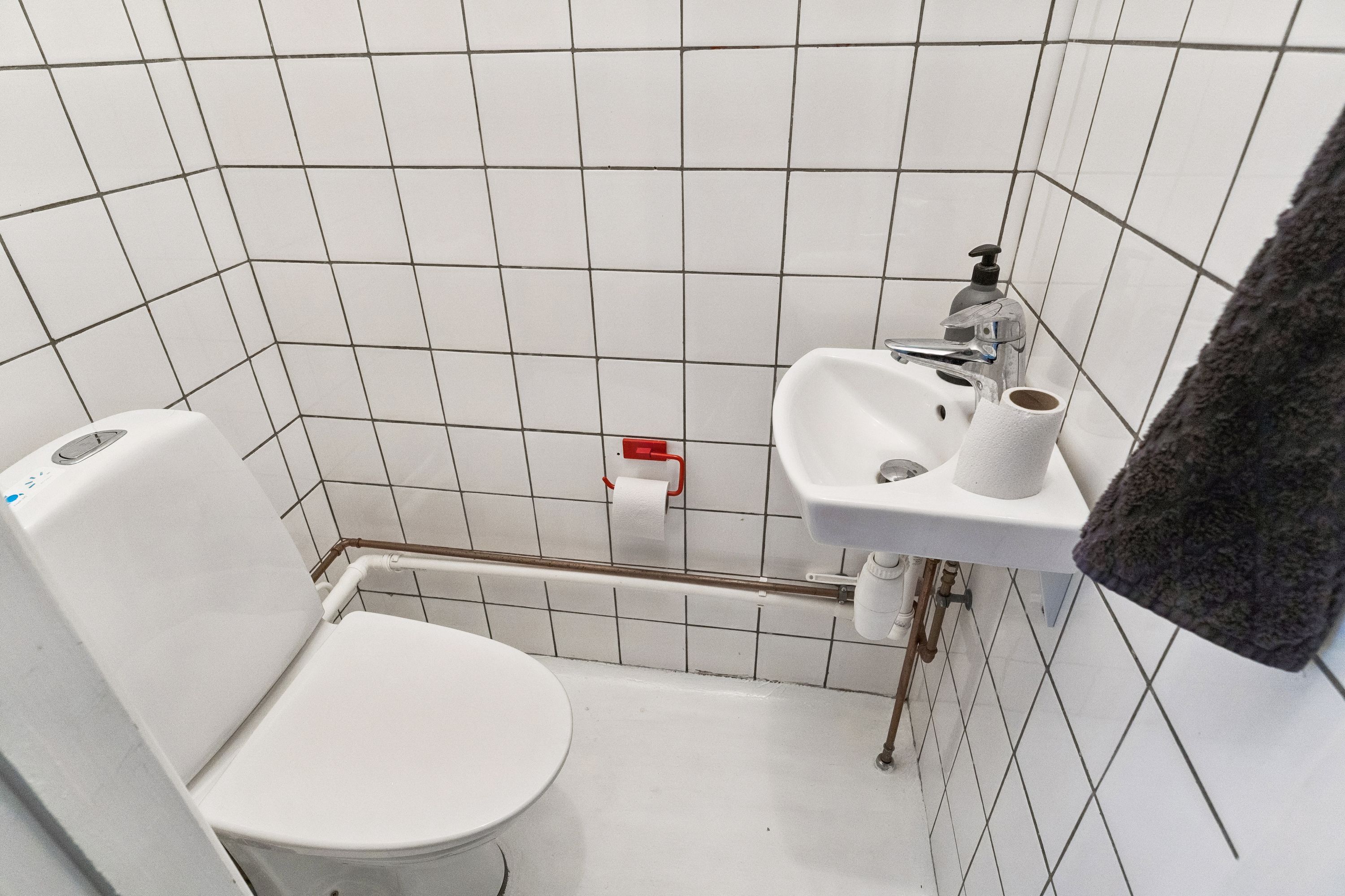 Toilette med hvidt toilet, håndvask, to toiletpapirruller og håndsæbe. Toiletpapirholder med toiletpapir og håndsæbeflaske på væggen.