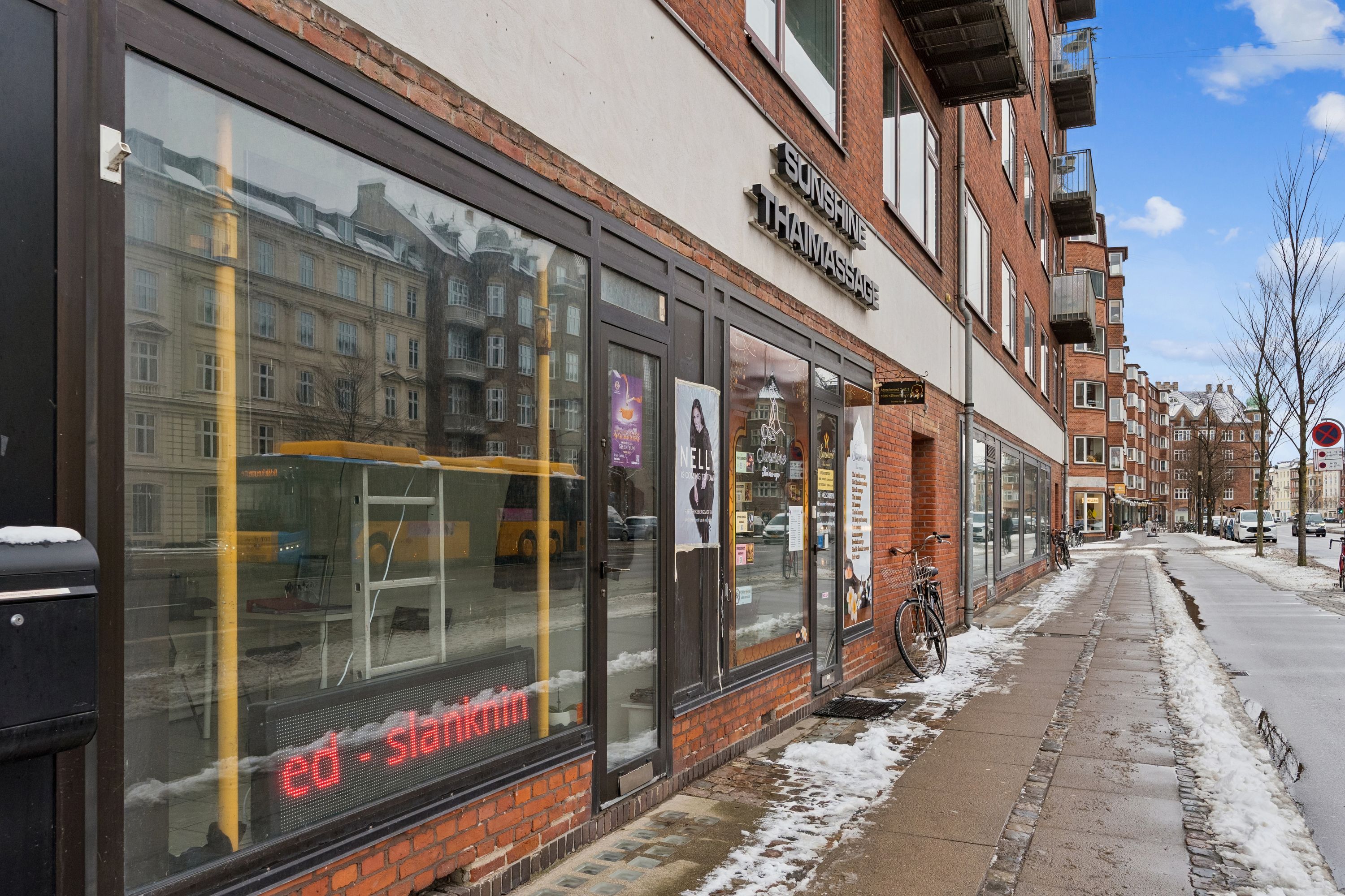 Butik med glassdøre og skilte, der læser "Sunshine Thaimassage" og "Ed-Slankmin". Sne dækker fortov og gaden med biler og cykler parkeret. Bygninger med røde mursten og mange vinduer langs vejen